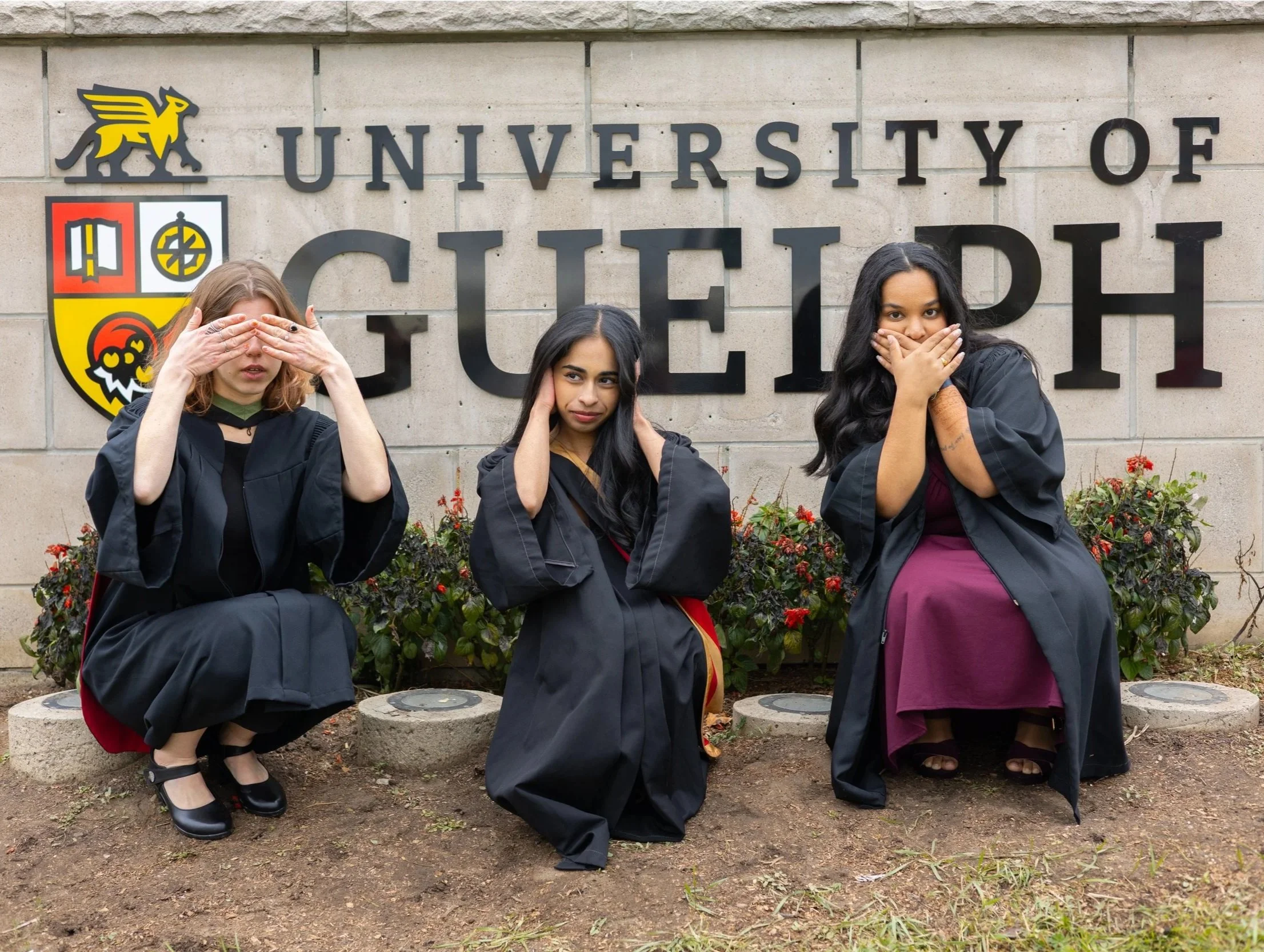 Three women in graduation gowns sitting on the ground in front of a sign that reads "University of Guelph". The woman on the left has light brown hair and is covering her eyes with her hands. The woman in the middle has long dark hair and is covering