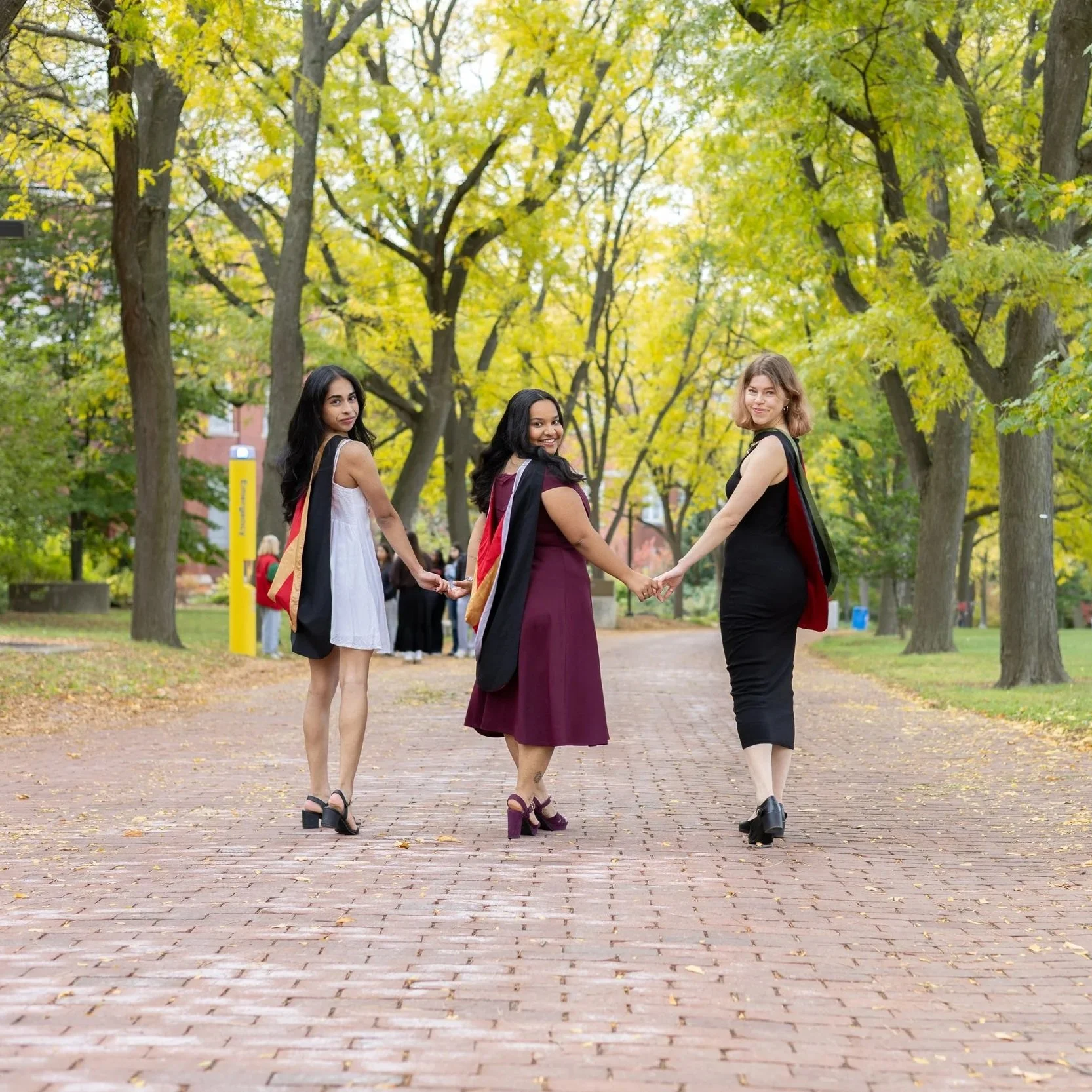 Three women in graduation gowns holding hands while walking in a park with trees and yellow leaves.