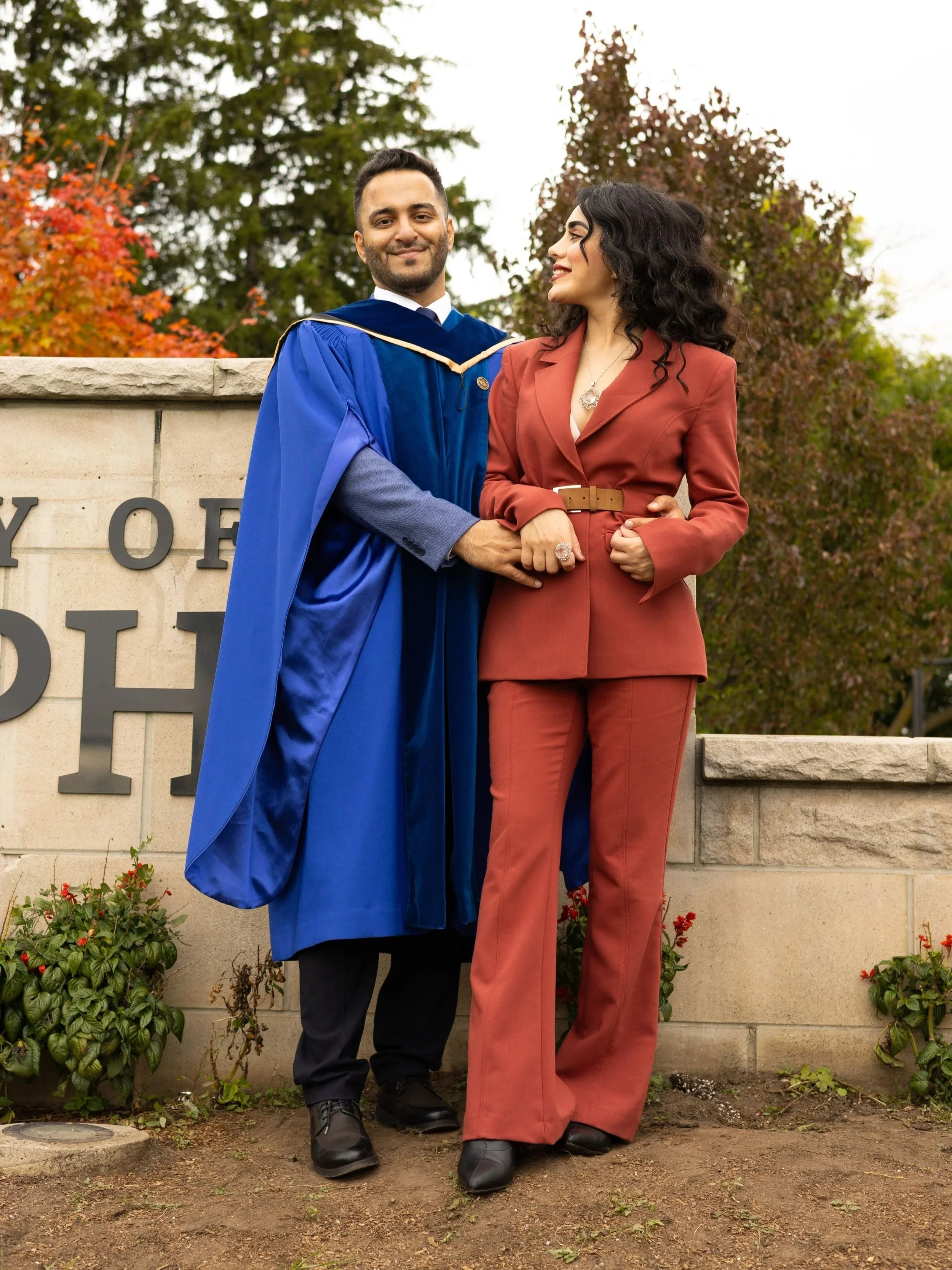 A man in a blue graduation gown and cap stands next to a woman in a rust-colored suit, holding her hand, next to a stone sign with part of it visible. They are outdoors with trees and an overcast sky in the background.