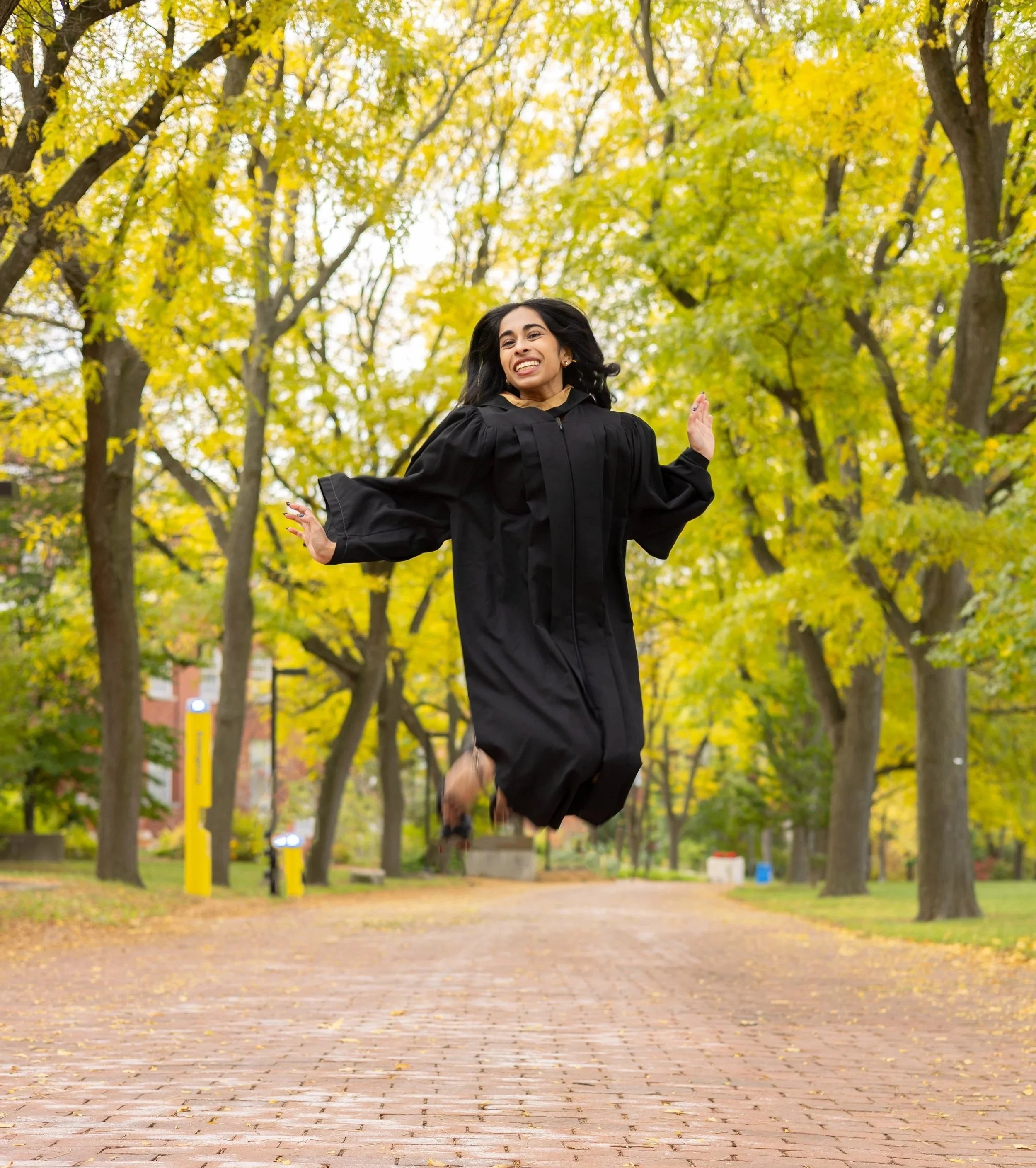 A woman in a black graduation gown jumping in a park with autumn yellow leaves.