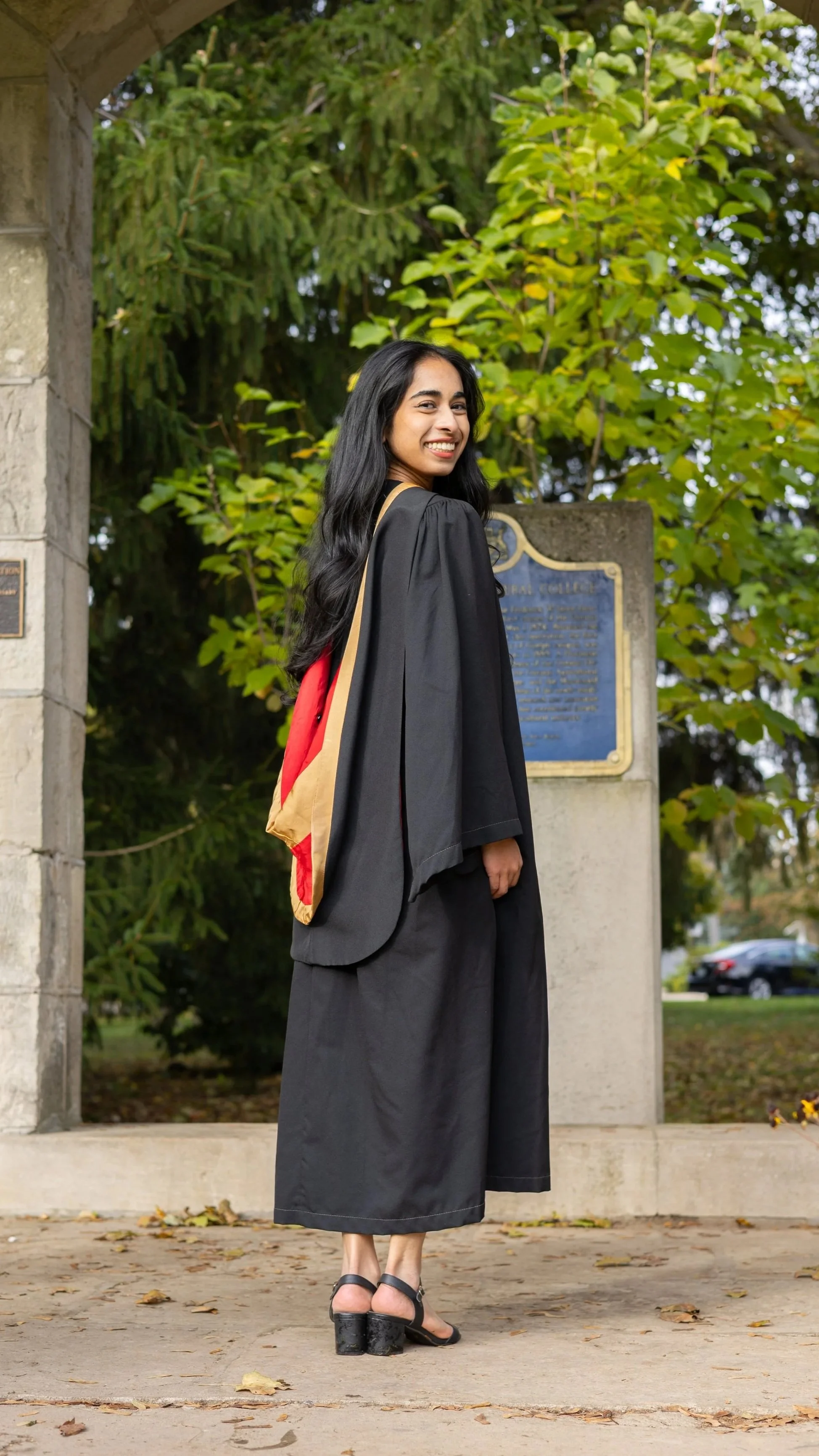 A young woman in a black graduation gown and cap, smiling, stands outdoors near a stone monument with a plaque, with green trees and cars in the background.