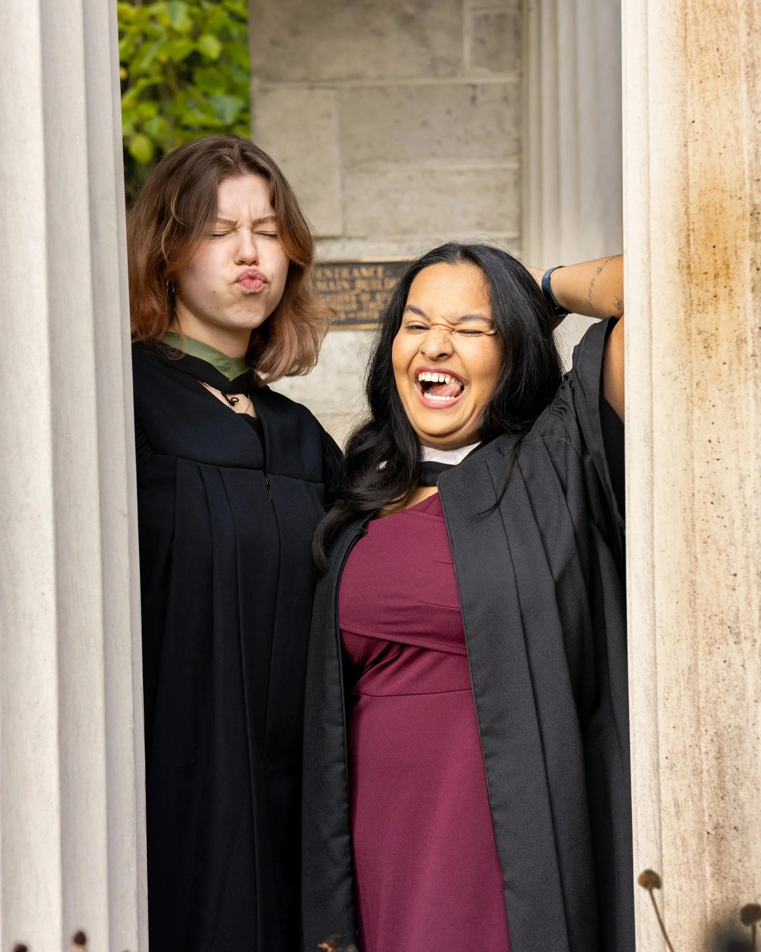 Two women in graduation gowns making funny faces outside a building.