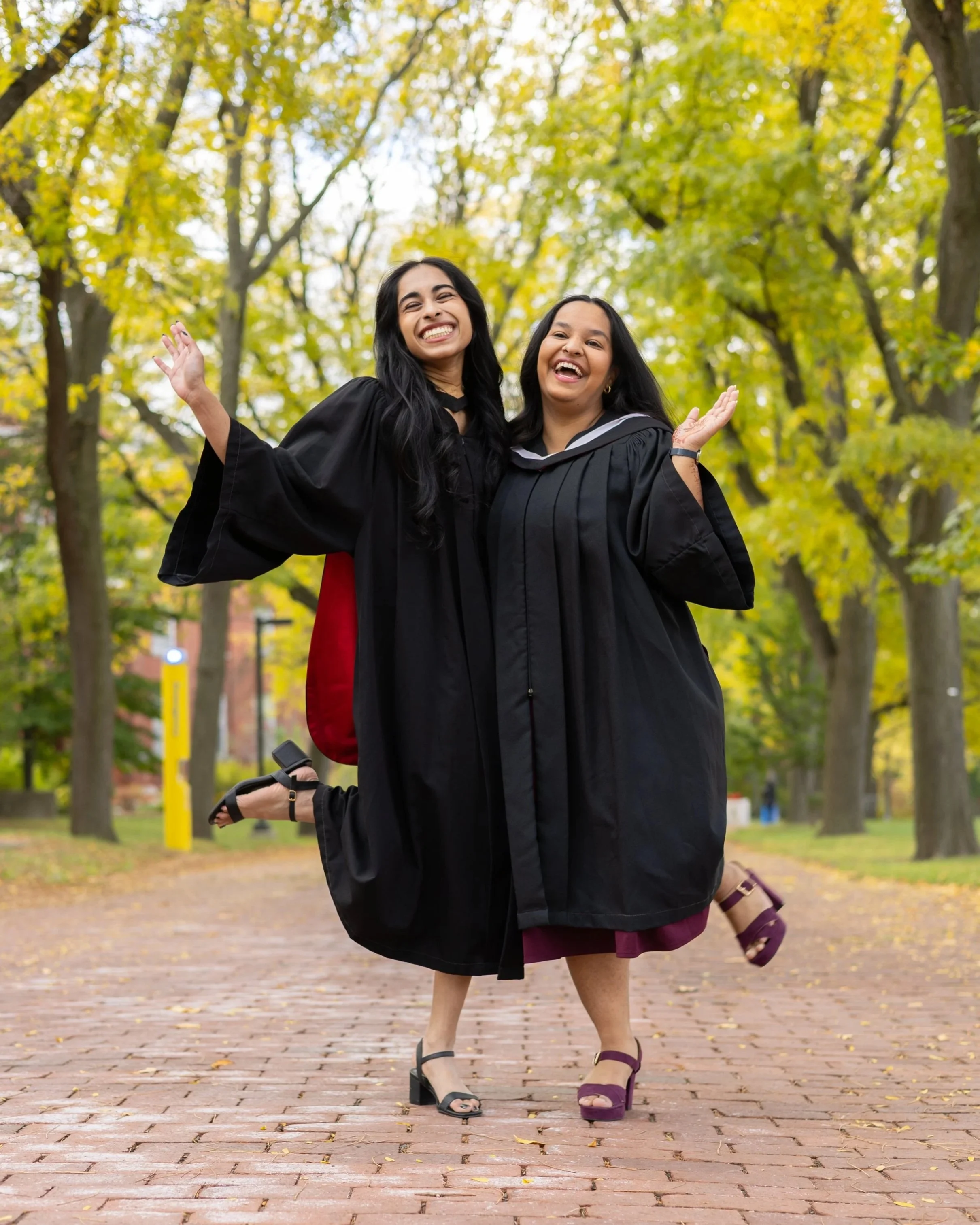 Two women in graduation caps and gowns celebrating, standing on a brick path in a park with trees and fall leaves.