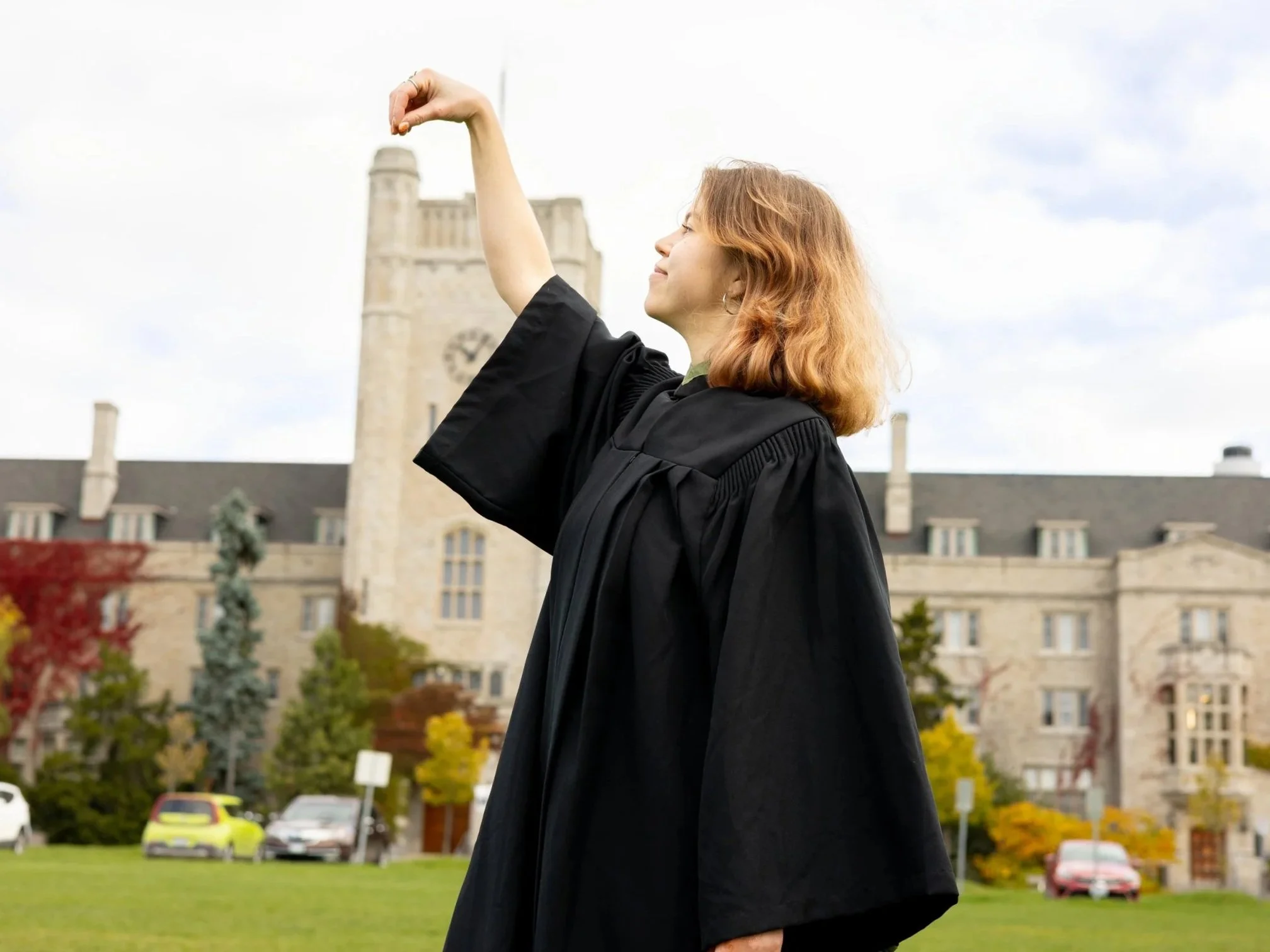 A woman in a graduation gown, standing outdoors in front of a historic building with a clock tower, appears to be celebrating her graduation.