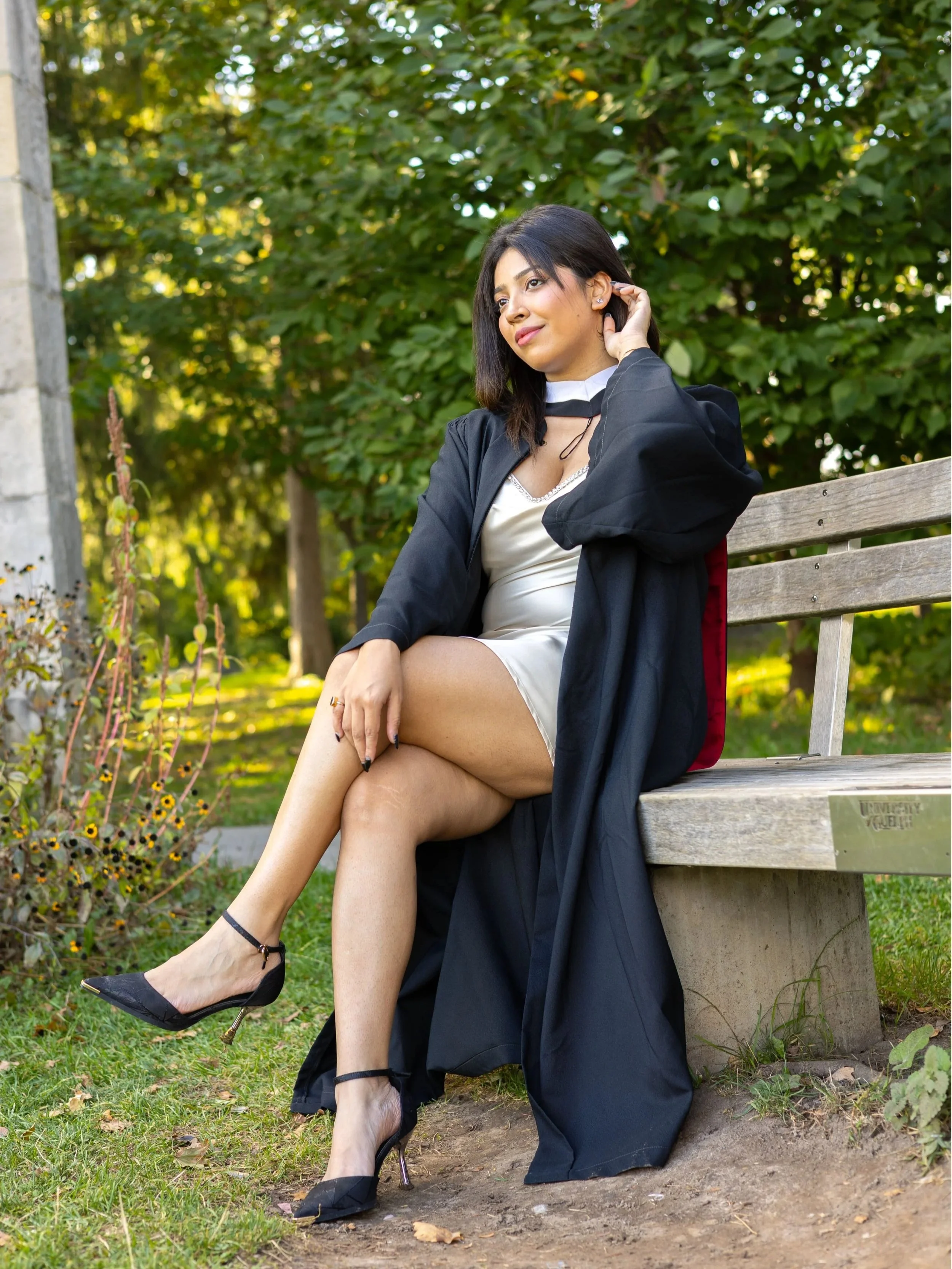 A woman sitting on a wooden park bench in a park, wearing a graduation gown with her legs crossed, black heels, a light-colored dress, and a graduation stole, with trees and bushes in the background.