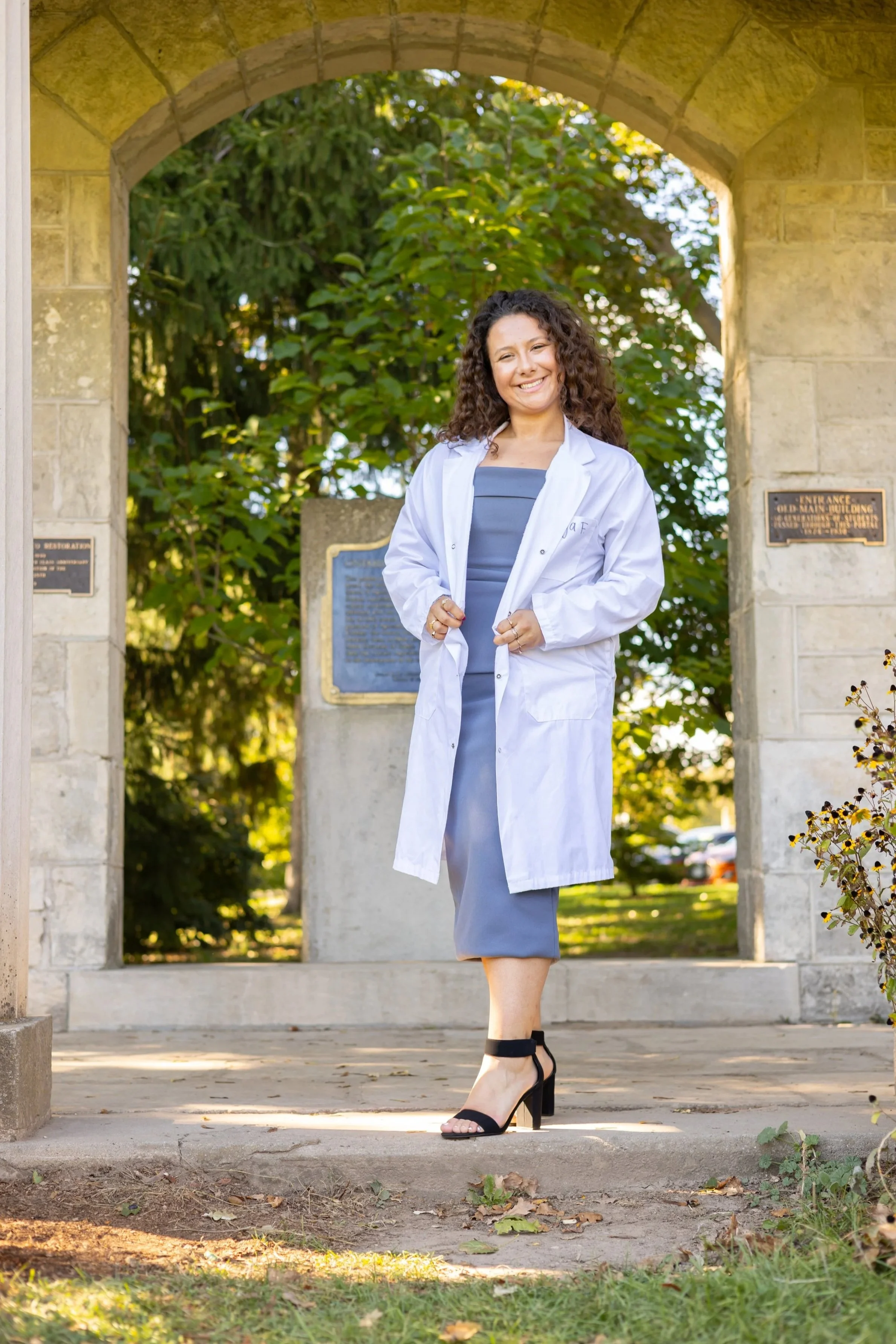 A woman with curly hair wearing a white lab coat, gray dress, and high heels standing under a stone archway outdoors, smiling at the camera with trees and plaques in the background.