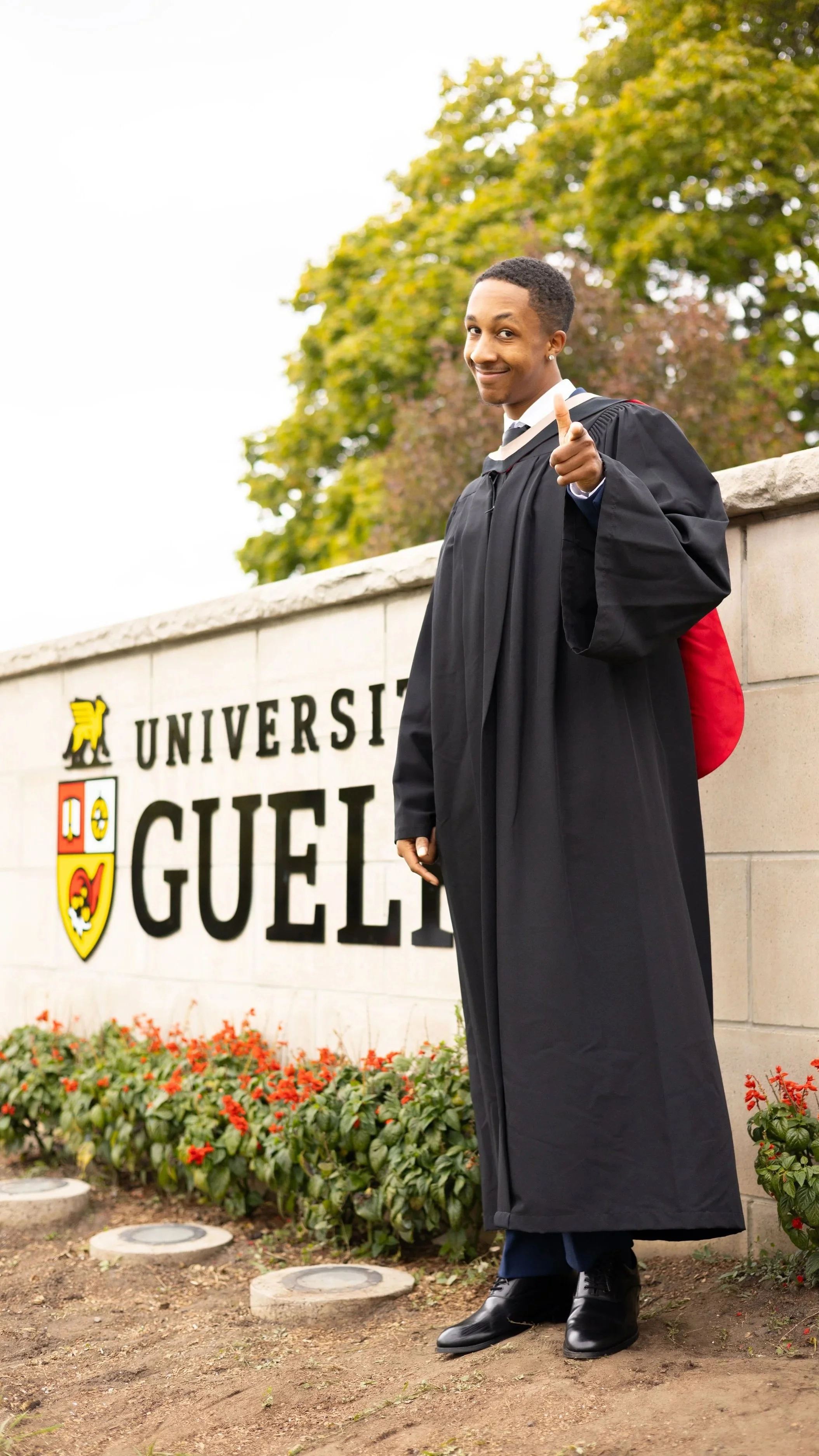 A graduate student in cap and gown standing in front of the University of Guelph sign, smiling and giving a thumbs up.