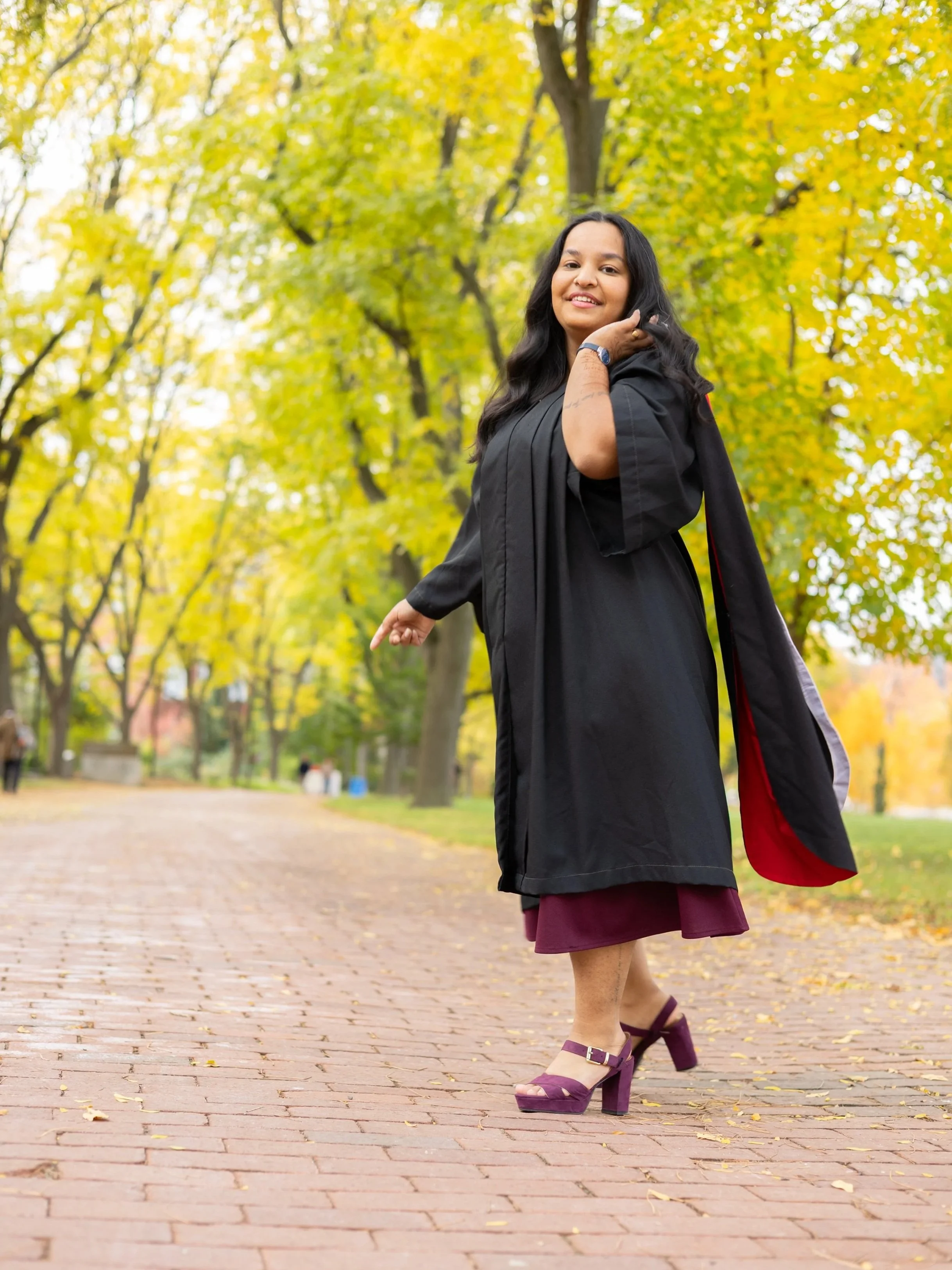 A woman in a graduation gown and heels standing on a brick path in a park with yellow autumn leaves.