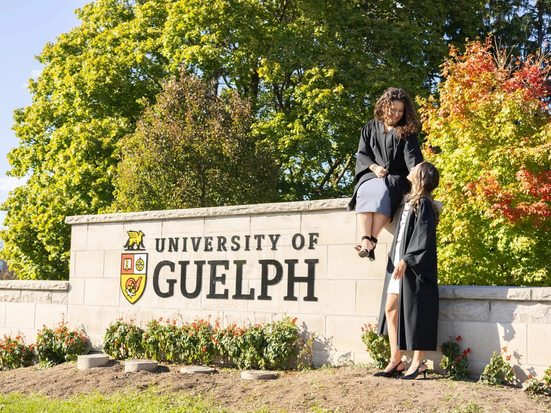 Two graduates in caps and gowns celebrate in front of a sign that reads 'University of Guelph' with colorful trees and flowers in the background.