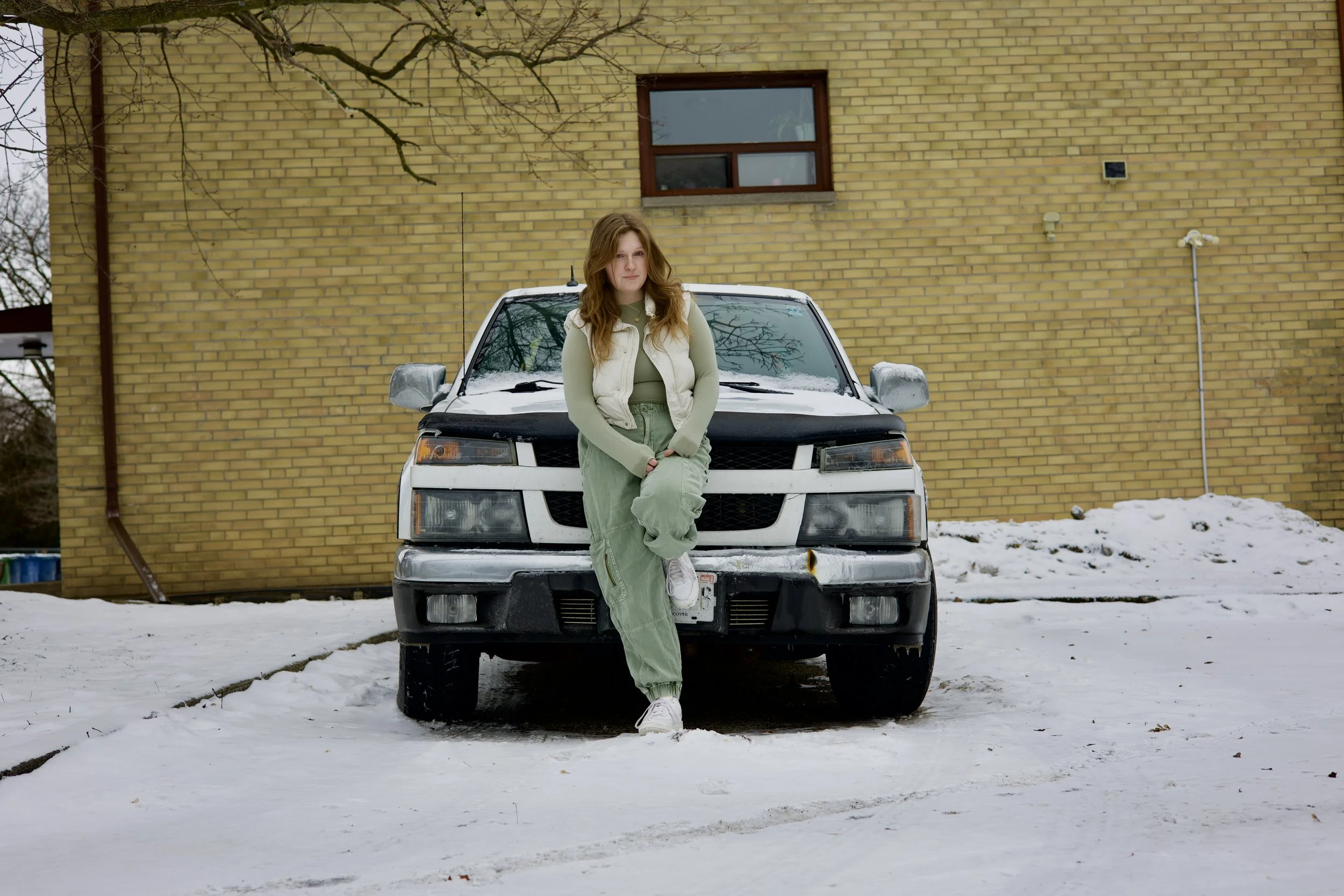 A young woman with long red hair, wearing a green sweatshirt, light green pants, white sneakers, and a beige vest, leaning against the front of a black and white SUV parked on snow in front of a yellow brick building with a single window.