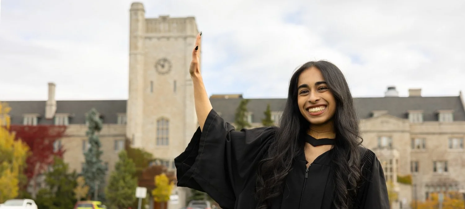 A young woman in graduation gown smiling and waving outside a historic building with autumn trees in the background.