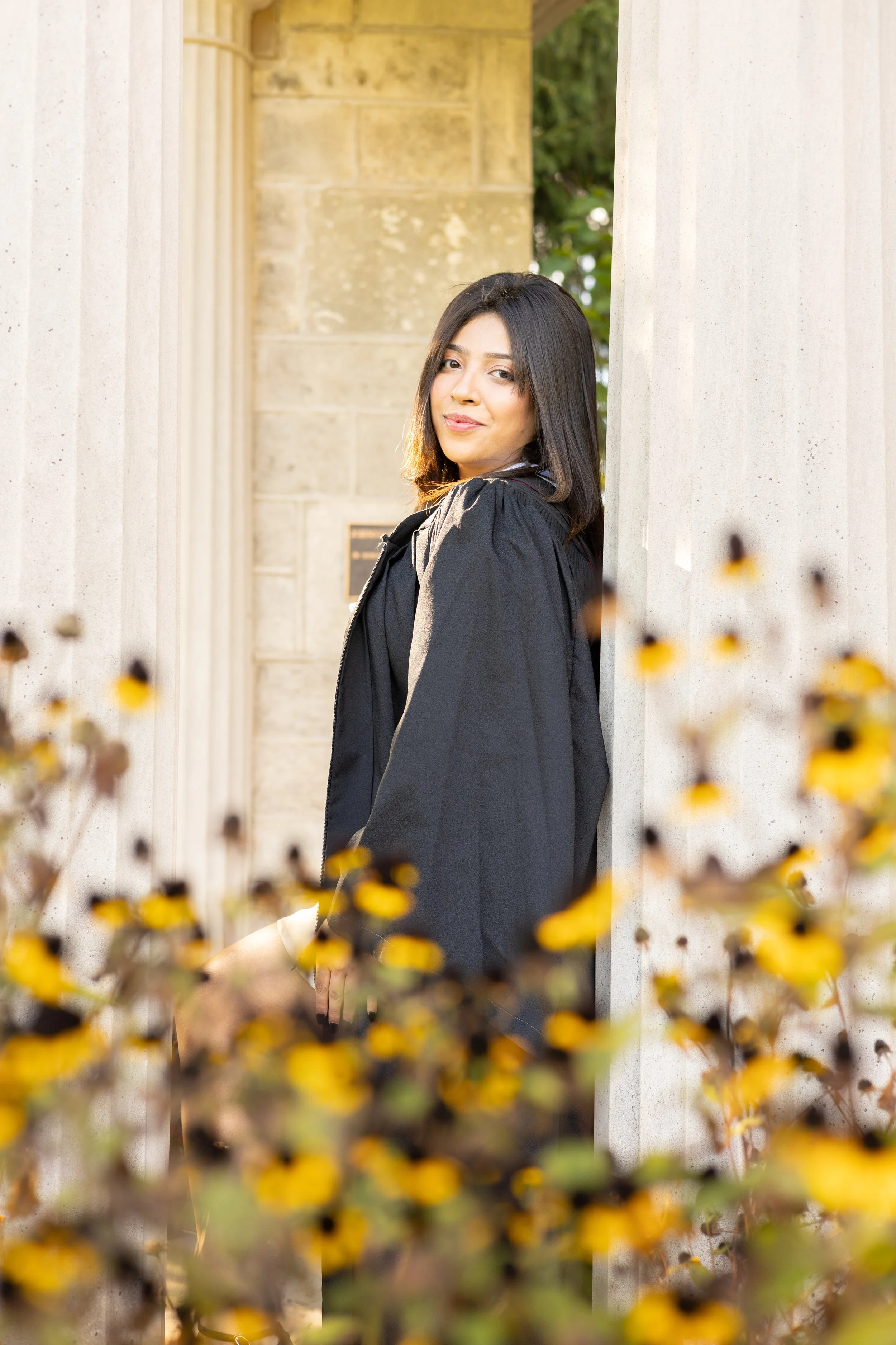 A young woman in a graduation gown leaning against a stone pillar, with yellow flowers in the foreground and trees in the background.
