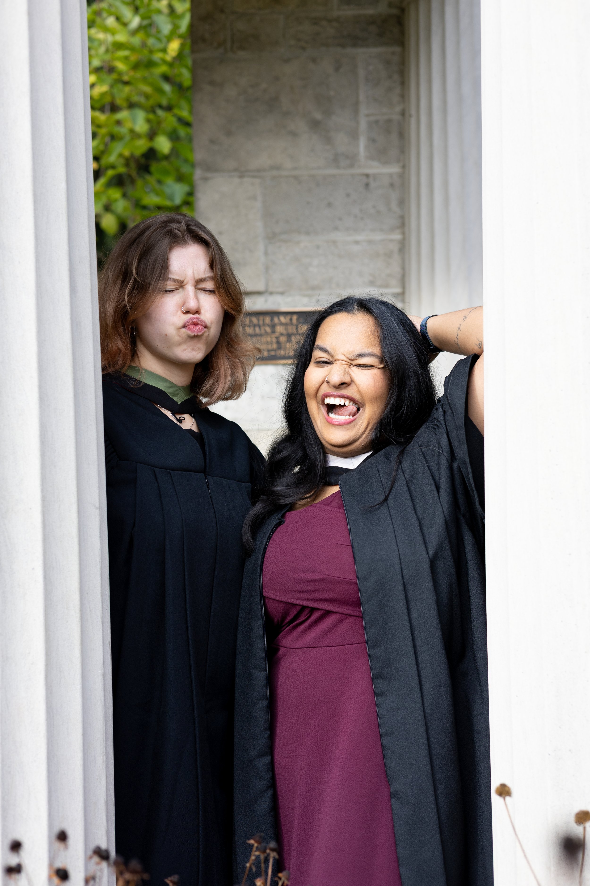 Two women in graduation gowns making funny faces, standing at a graduation ceremony outside a building.
