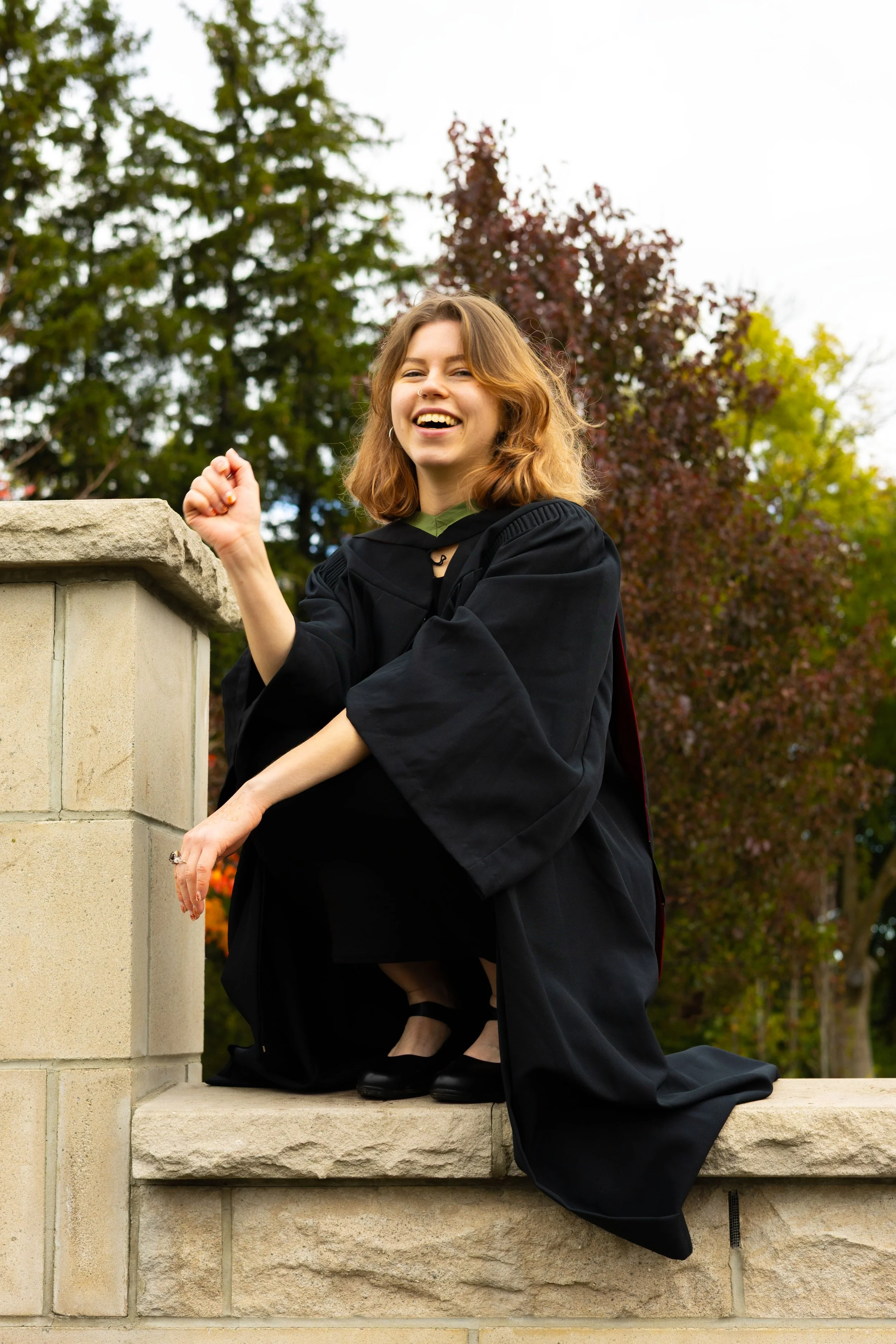 A young woman in a graduation gown and cap sitting on a stone ledge outdoors, smiling and looking happy with trees and sky in the background.