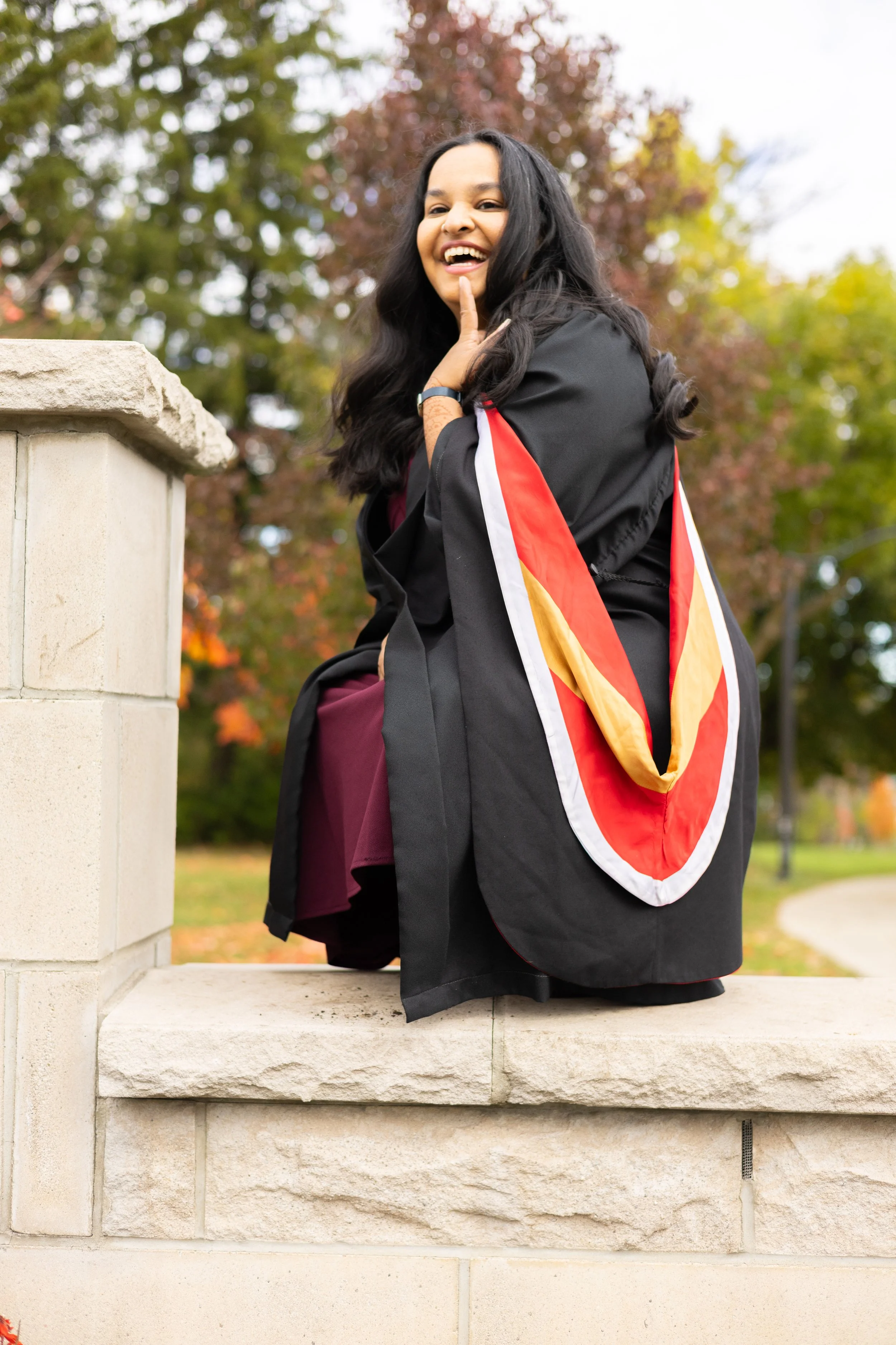 A woman in a graduation gown sitting on a stone wall outdoors, smiling and touching her face, with colorful autumn trees in the background.