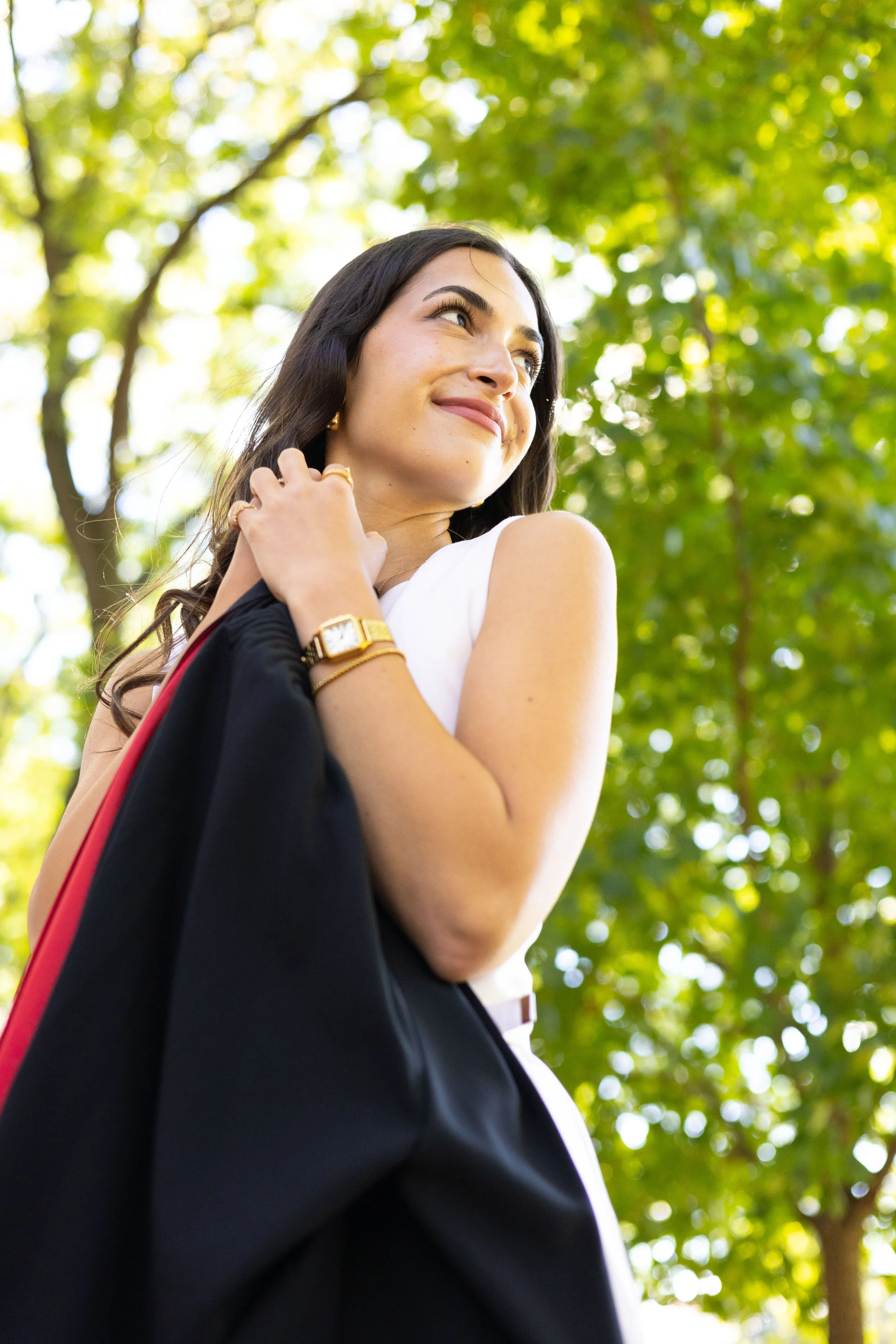 A young woman with dark hair smiling outdoors, holding a black garment with red lining over her shoulder, surrounded by green trees.
