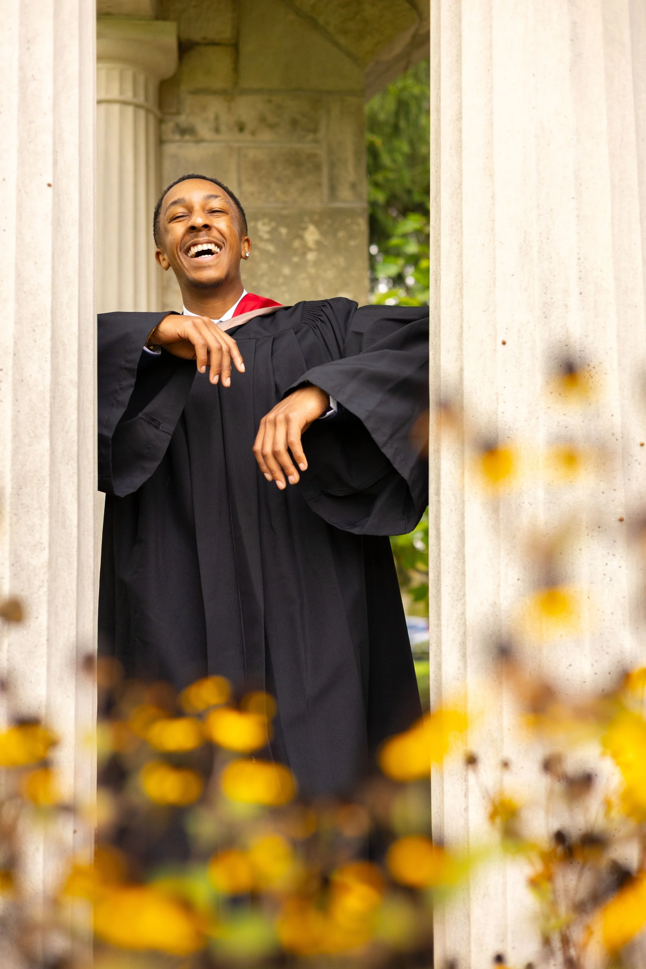 A young man in graduation gown and cap smiling and leaning on the stone frame of a building.