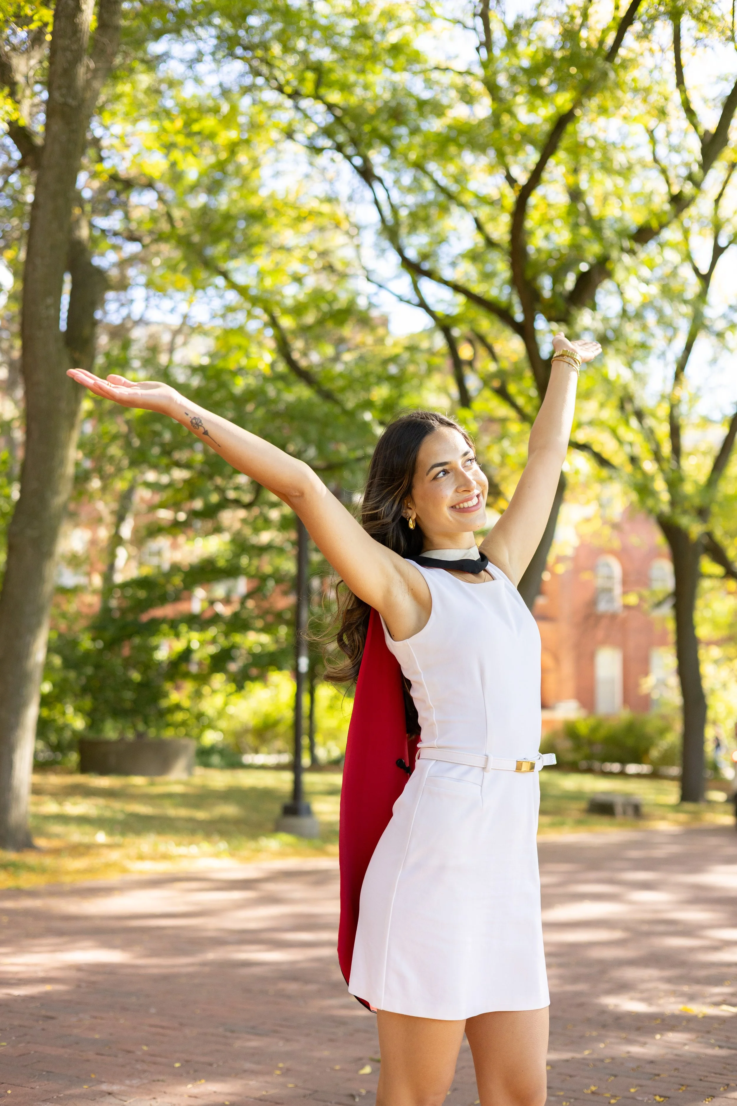 A woman in a white dress with a red cape standing outdoors on a sunny day, smiling with her arms raised in a joyful pose, surrounded by green trees and a brick building in the background.