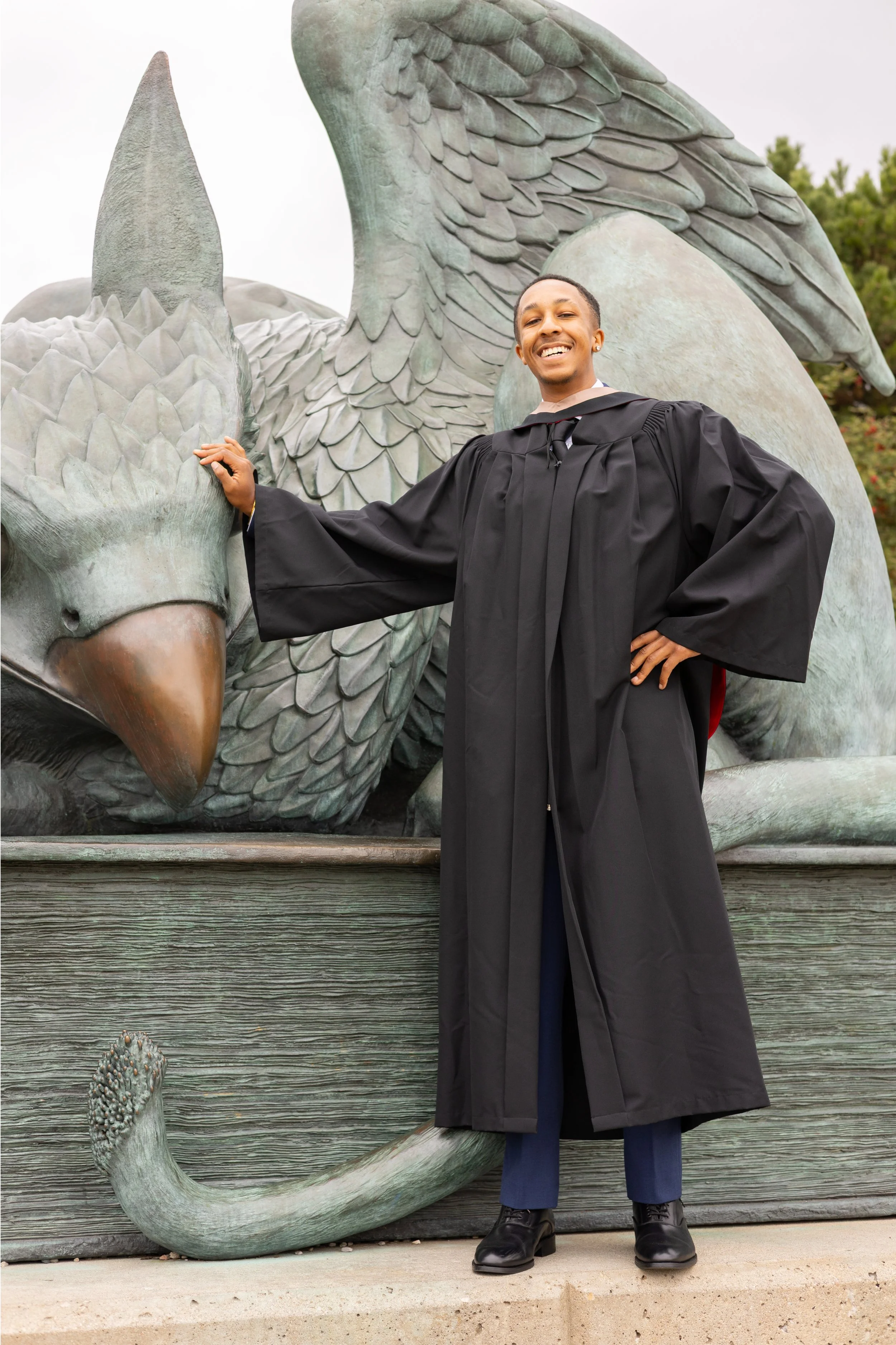A person in a black graduation gown and cap standing in front of a peacock statue, smiling and posing with one hand on the statue's head.