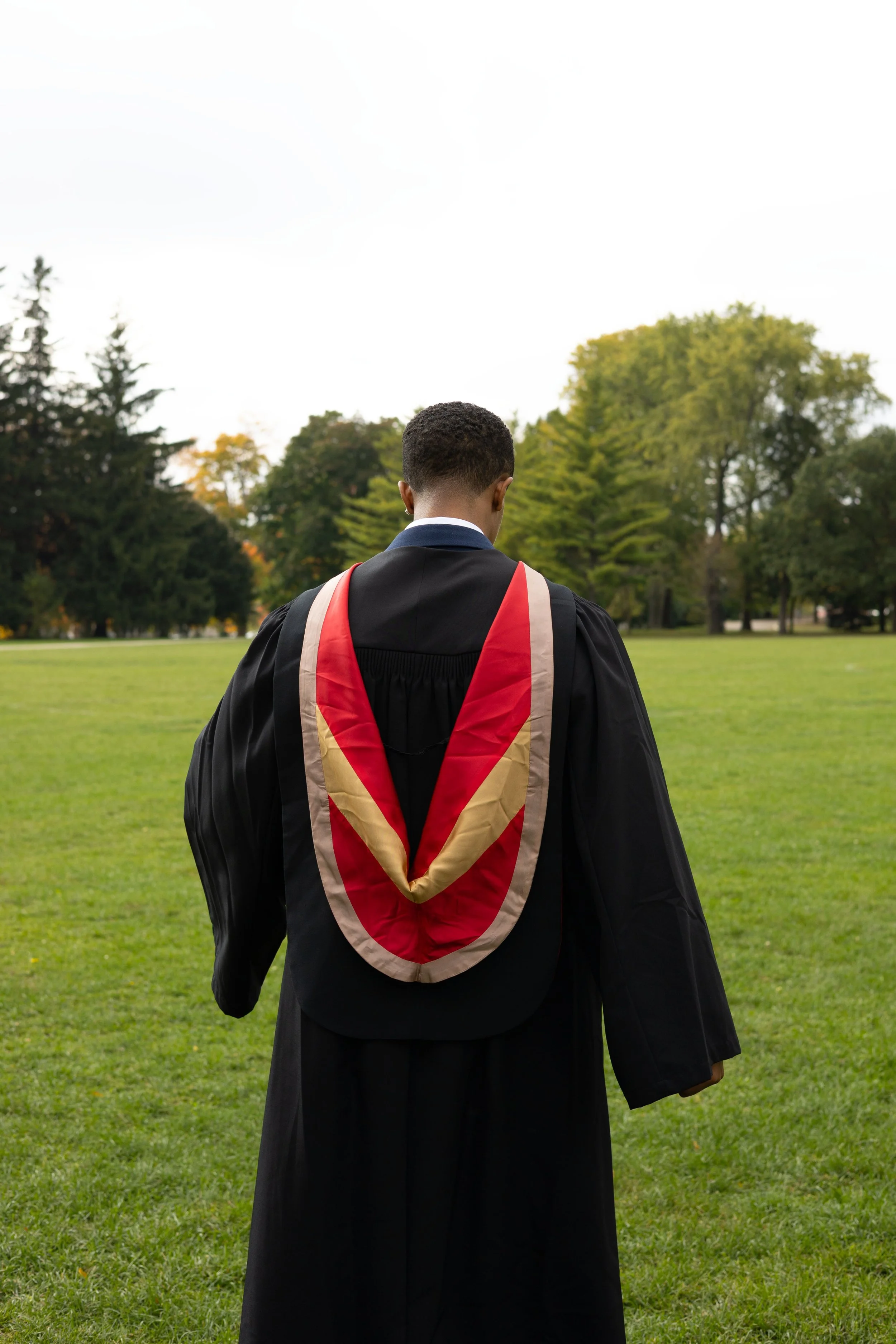 Graduation graduate in gown and cap with a red and beige hood, standing on a grassy field with trees in the background.
