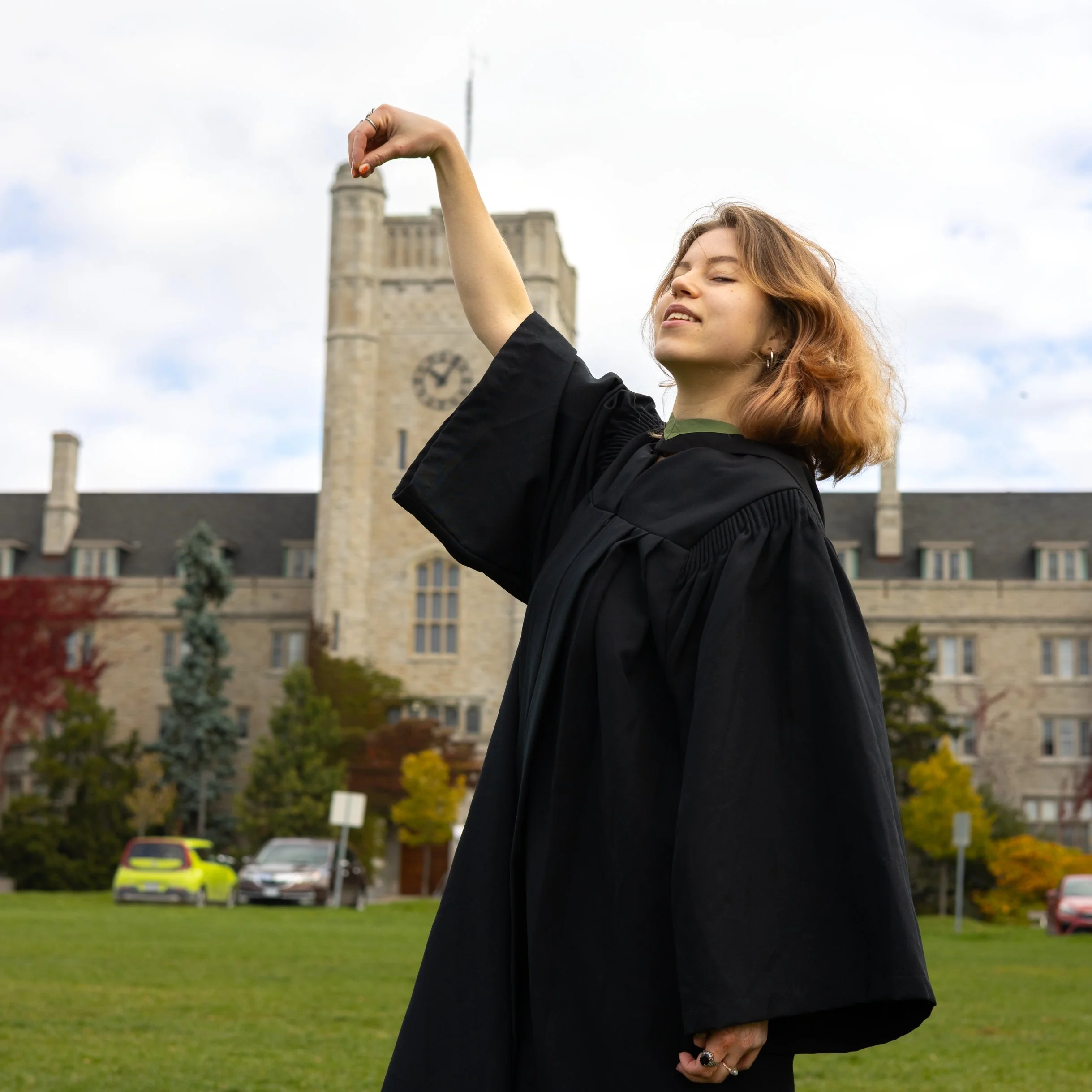 A young woman in a black graduation gown celebrating outdoors on a graduation day, with a historic building and parked cars in the background.
