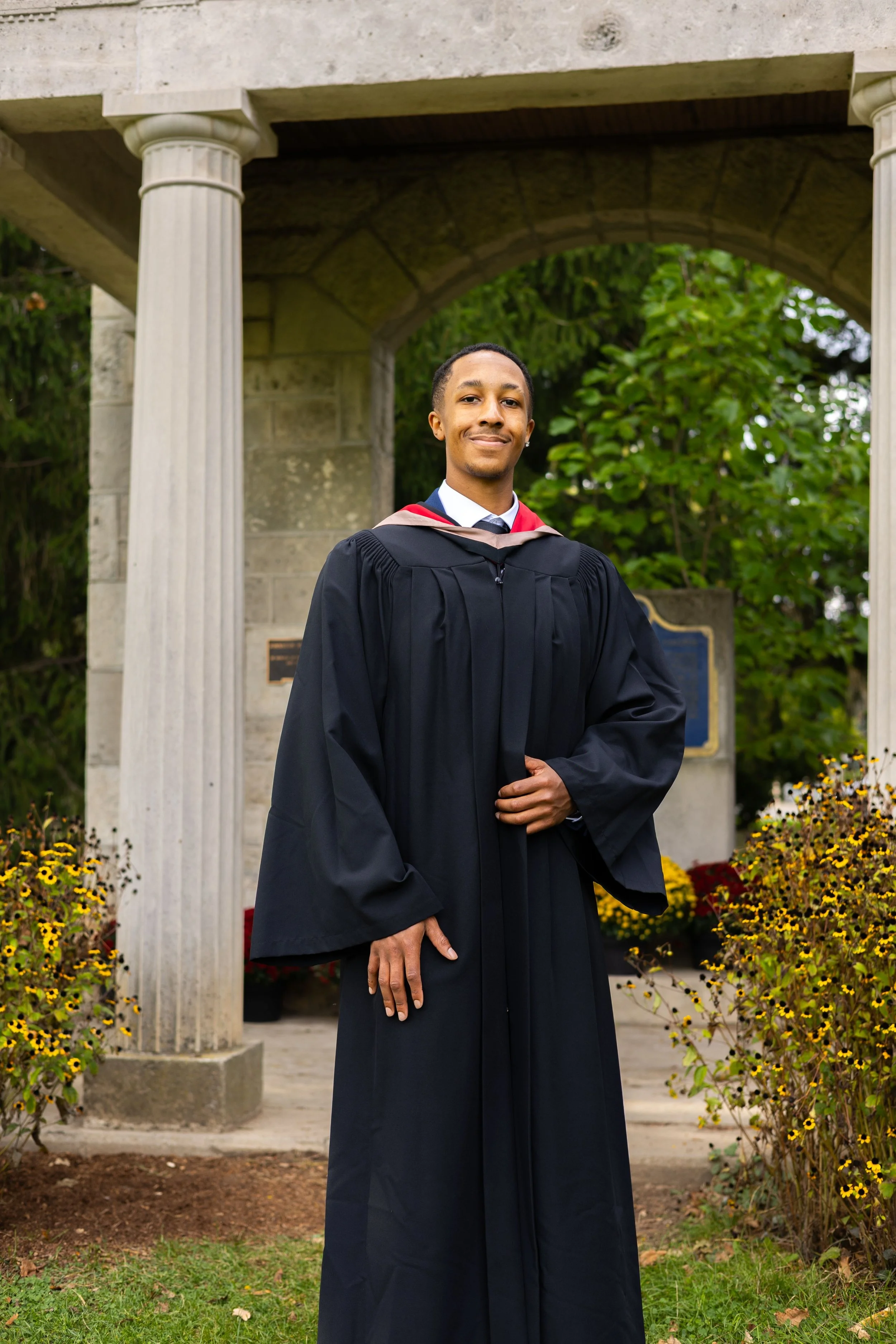 A young man in a graduation gown and cap standing outdoors in front of a stone structure with columns, surrounded by yellow flowers and greenery.