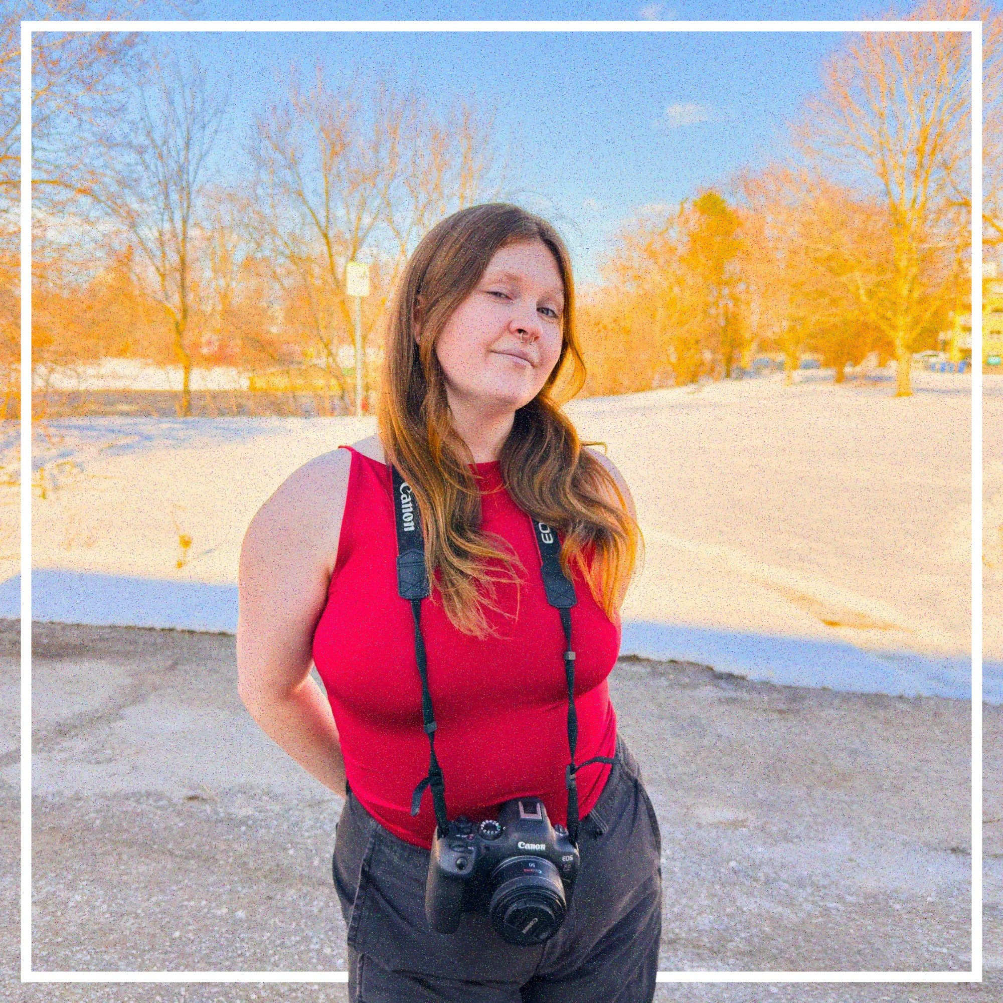 A young woman with long brown hair, wearing a red sleeveless top, standing outdoors on a sidewalk with trees and snow in the background, with a camera hanging around her neck.