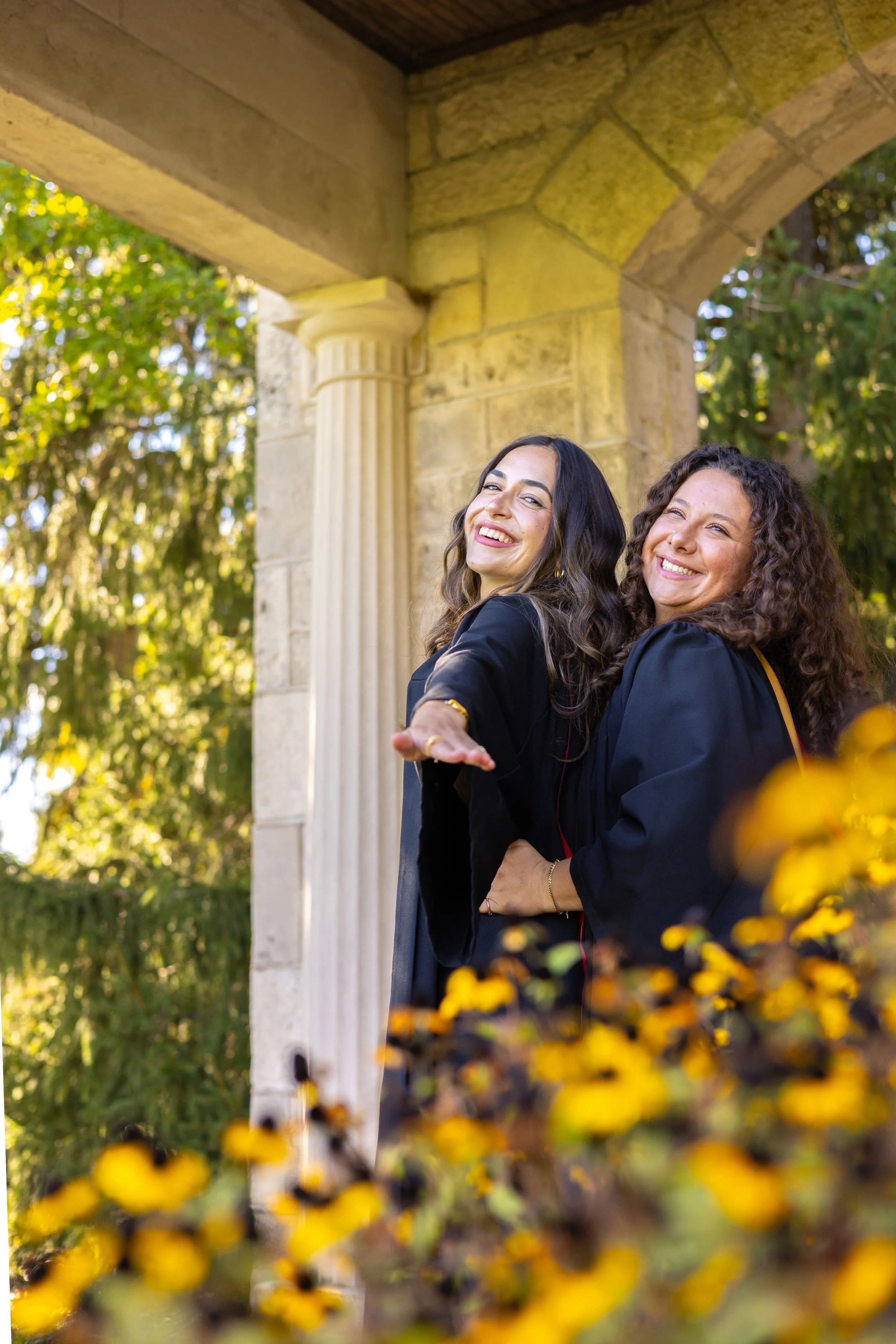 Two women in graduation robes celebrate outdoors under a stone archway with yellow flowers in the foreground.