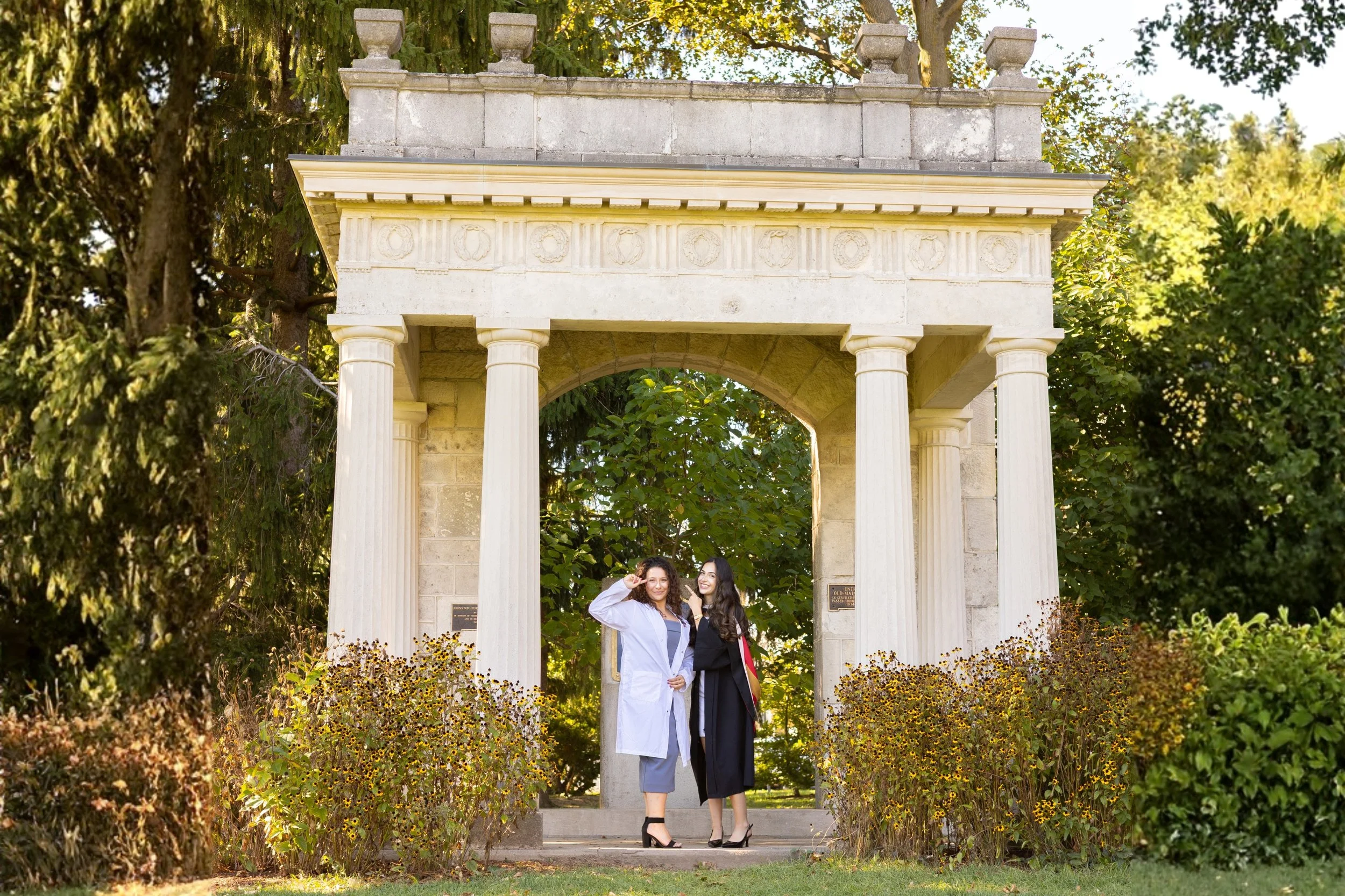 Two women in graduation attire standing under a stone archway with columns, surrounded by greenery and flowers.