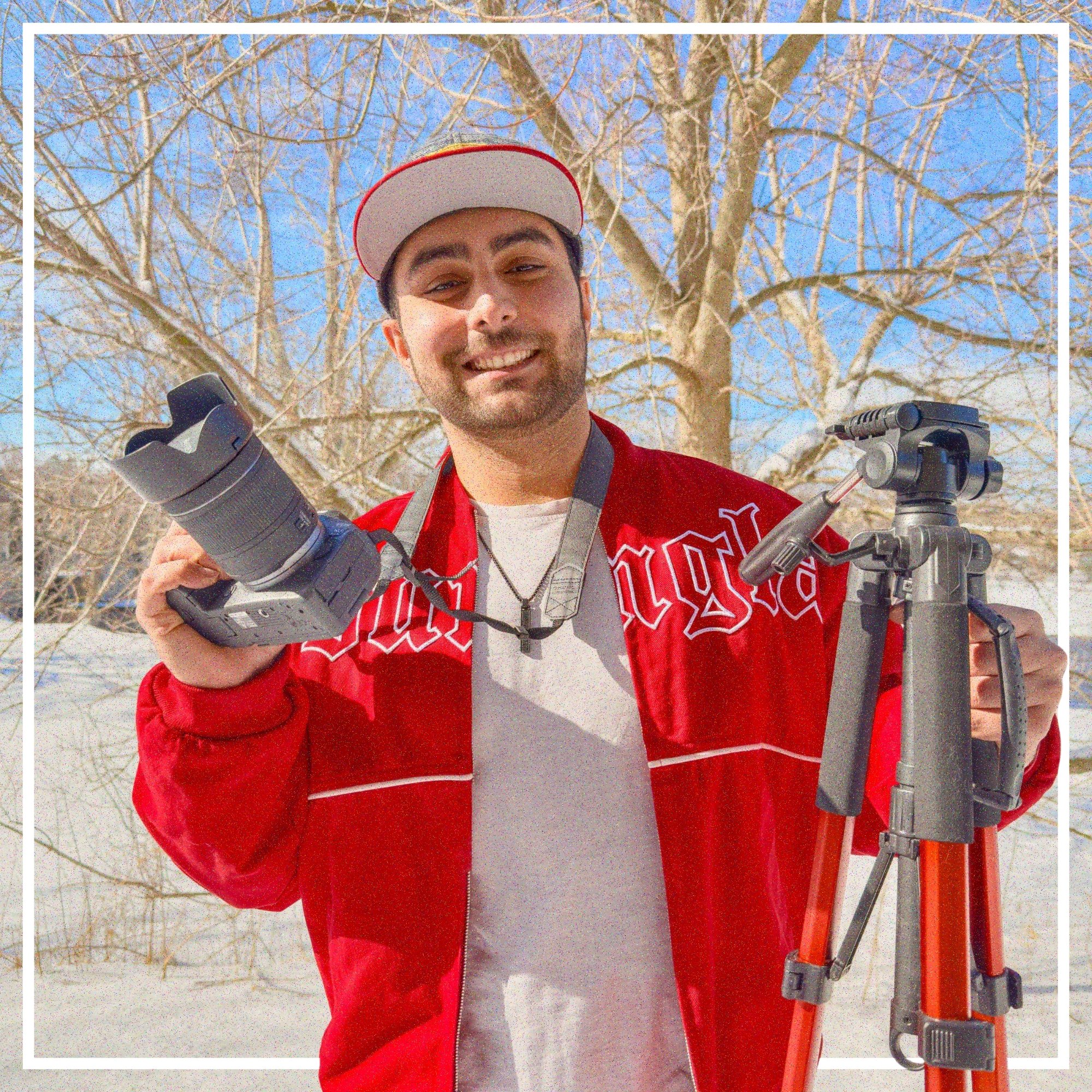 A man standing outdoors in a snowy landscape, holding a camera in his right hand and a tripod in his left, smiling at the camera.