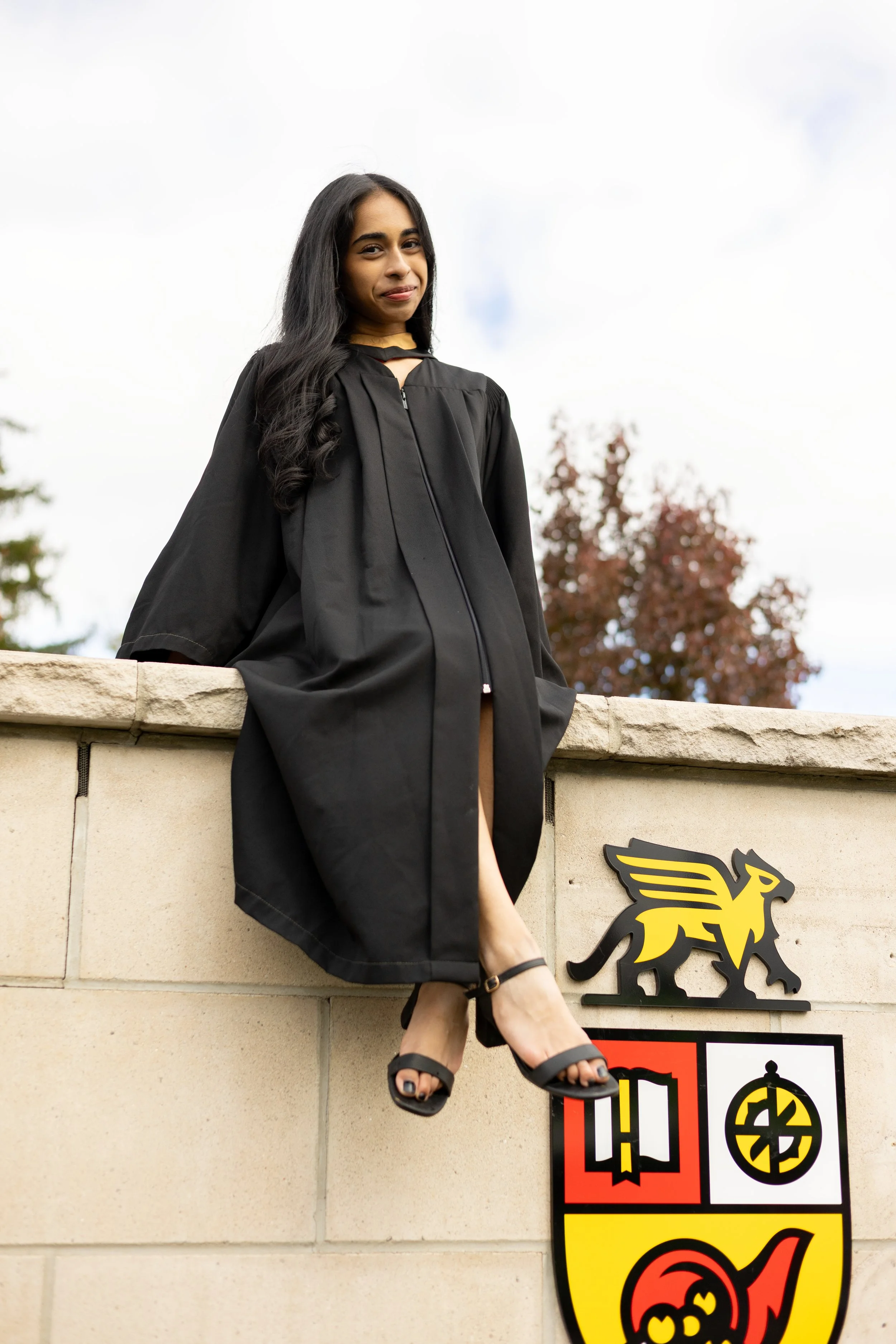 A young woman in a graduation gown sitting on a stone wall with university crest, outdoors with cloudy sky and trees in background.