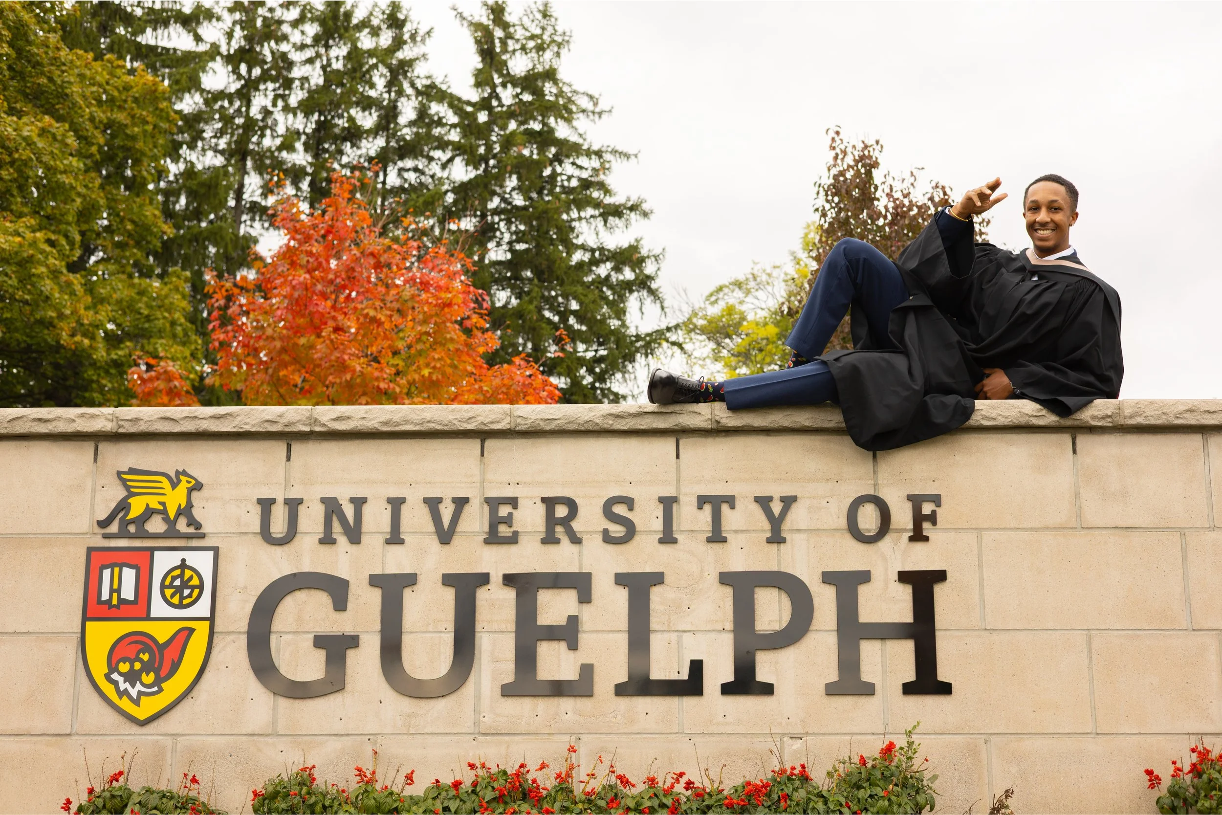 Graduate in black gown and cap lying on top of a stone wall with the University of Guelph sign, colorful autumn trees in the background.