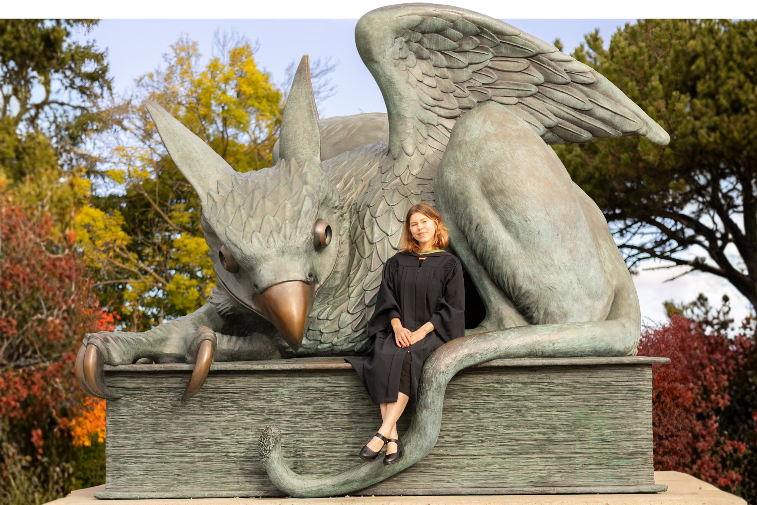 A woman in a black dress sitting on the edge of a large dragon sculpture, which has wings, horns, and claws, outdoors with trees and autumn foliage in the background.