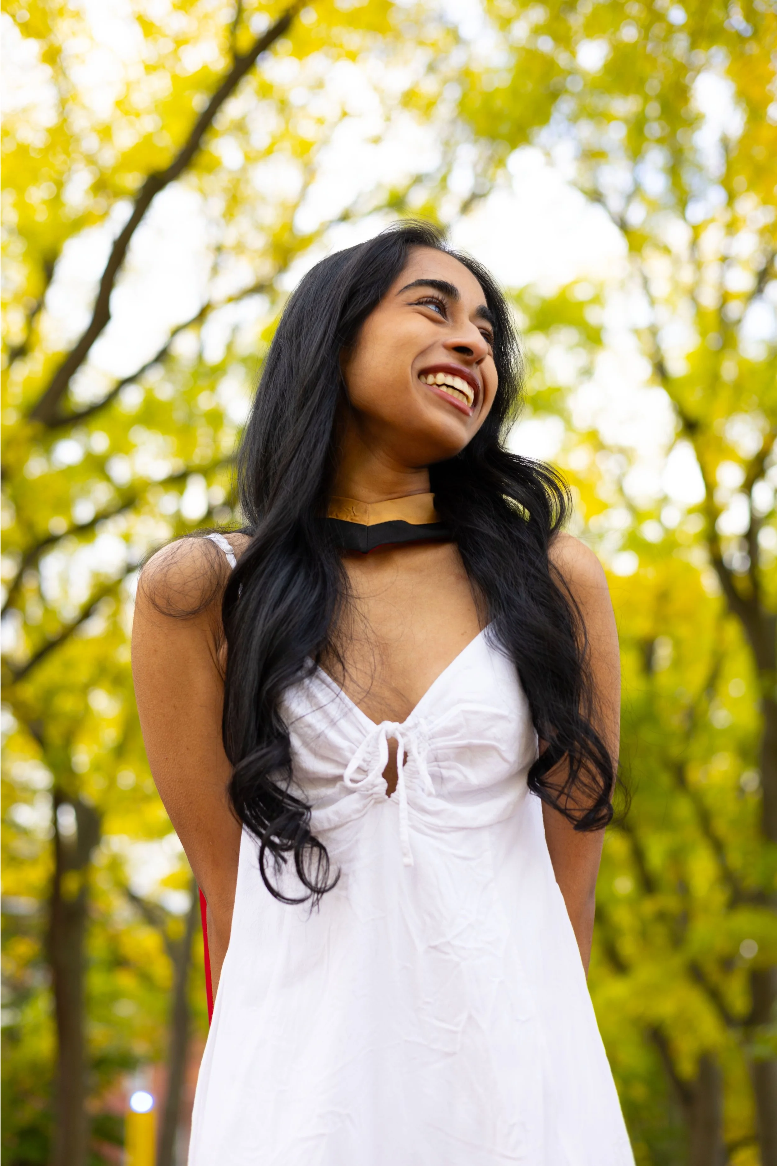 A young woman with long black hair smiling outdoors during fall, wearing a white dress and a black and yellow graduation stole.