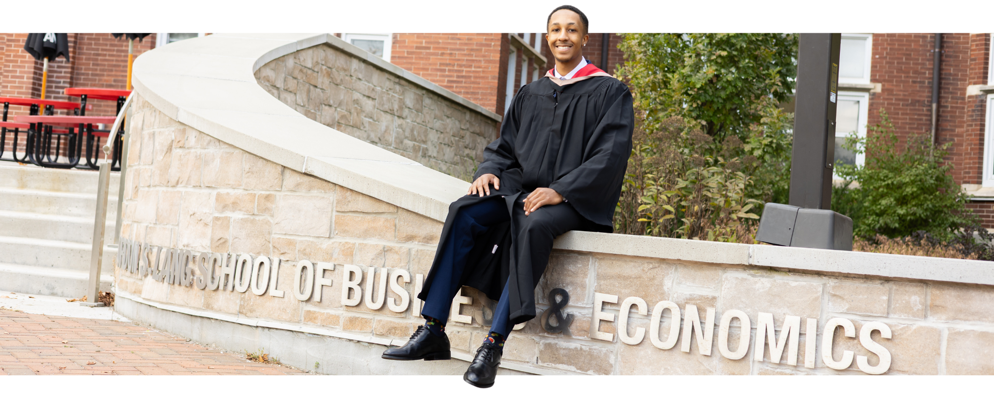 A young man in a black graduation gown and cap sitting on a low stone wall outside a building with a sign that reads 'Highland School of Business & Economics,' smiling at the camera.