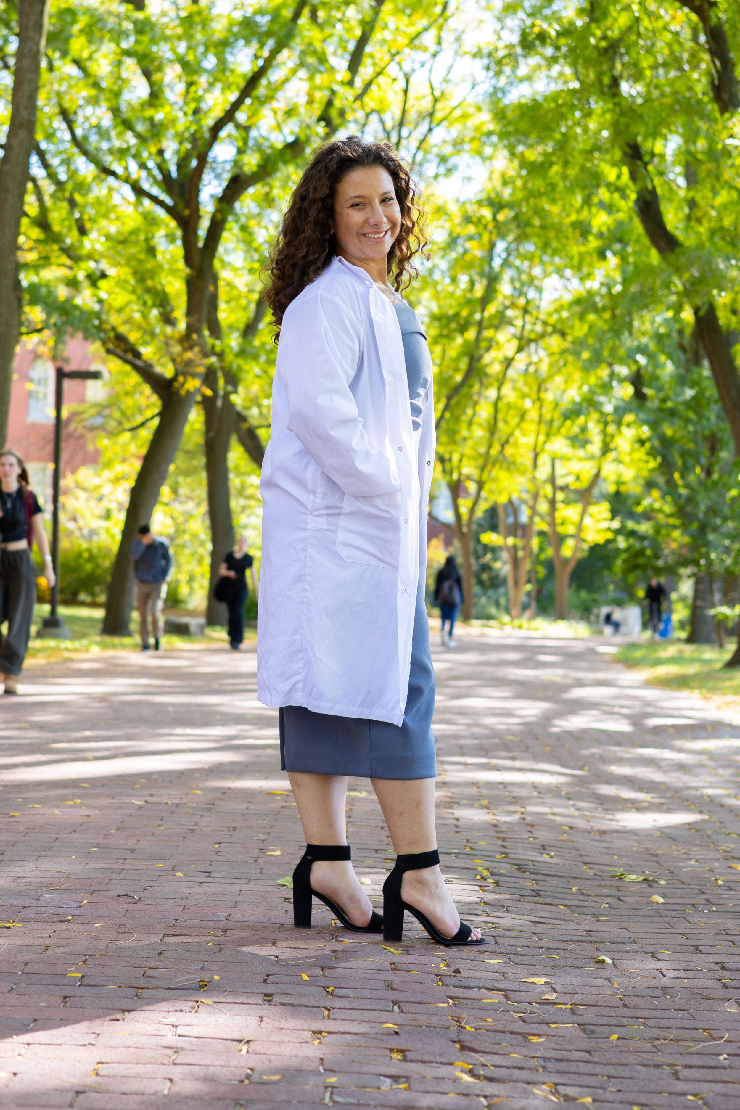 A woman with curly hair in a white coat posing outdoors on a brick pathway surrounded by green trees, with other people walking in the background.