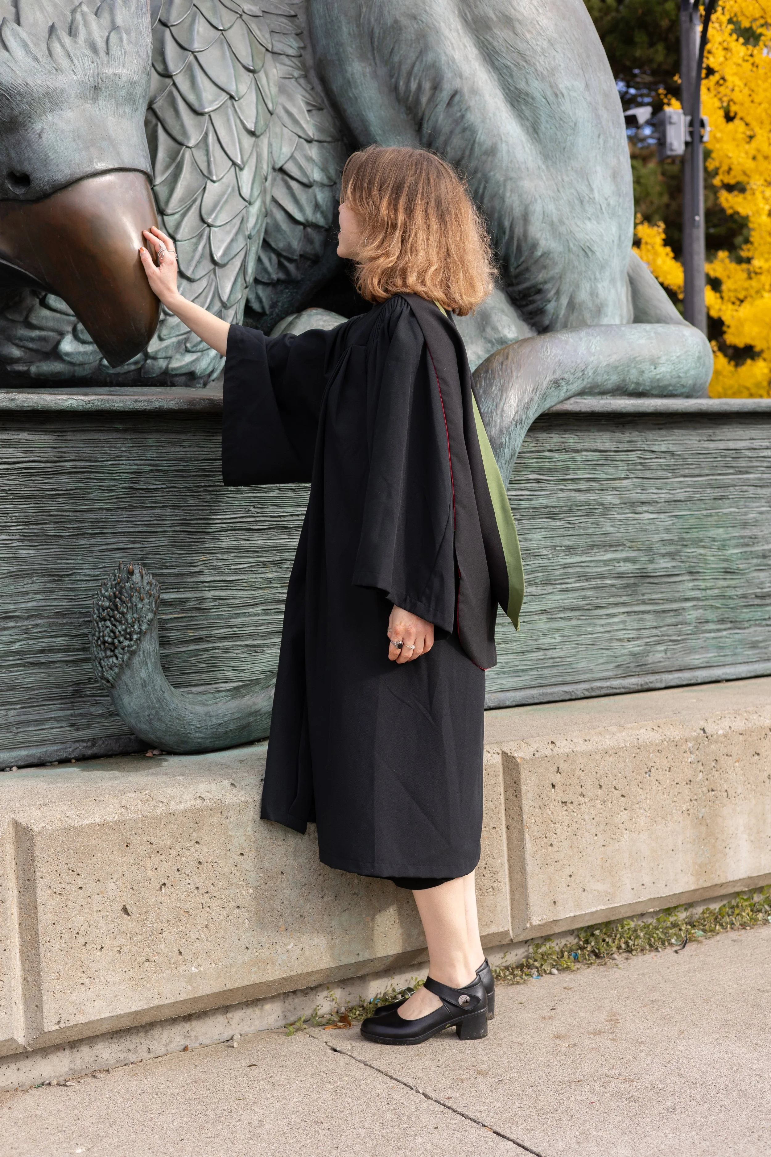 A woman in a black graduation gown touching the snout of a large bronze or metal sculpture of a dragon. The sculpture includes detailed scales and claws, and is set outdoors with trees and yellow leaves in the background.