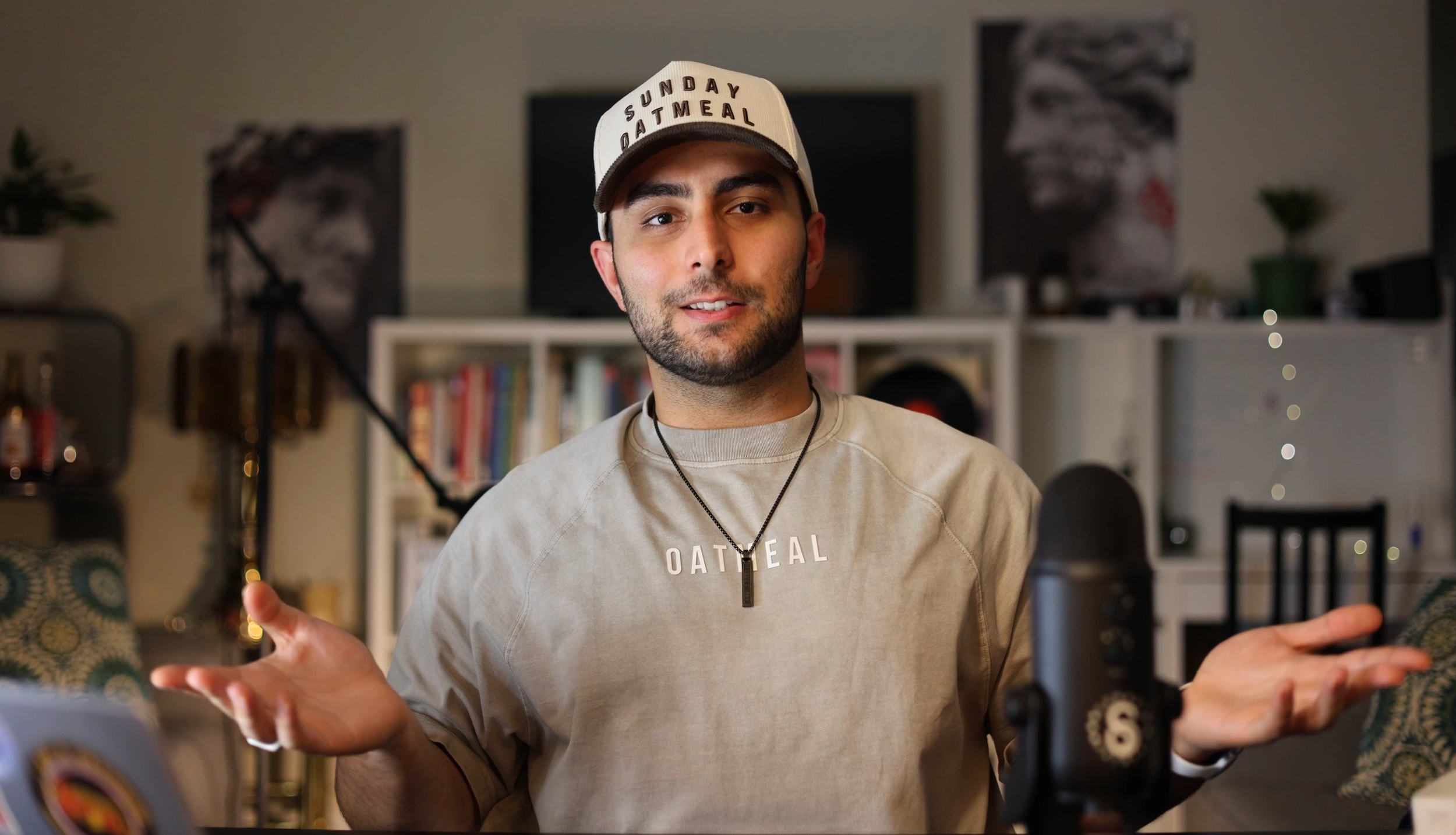 A young man with a beard and wearing a baseball cap that says 'Sunday Oatmeal', standing in a living room with a microphone in front of him, gesturing with his hands, and looking at the camera.