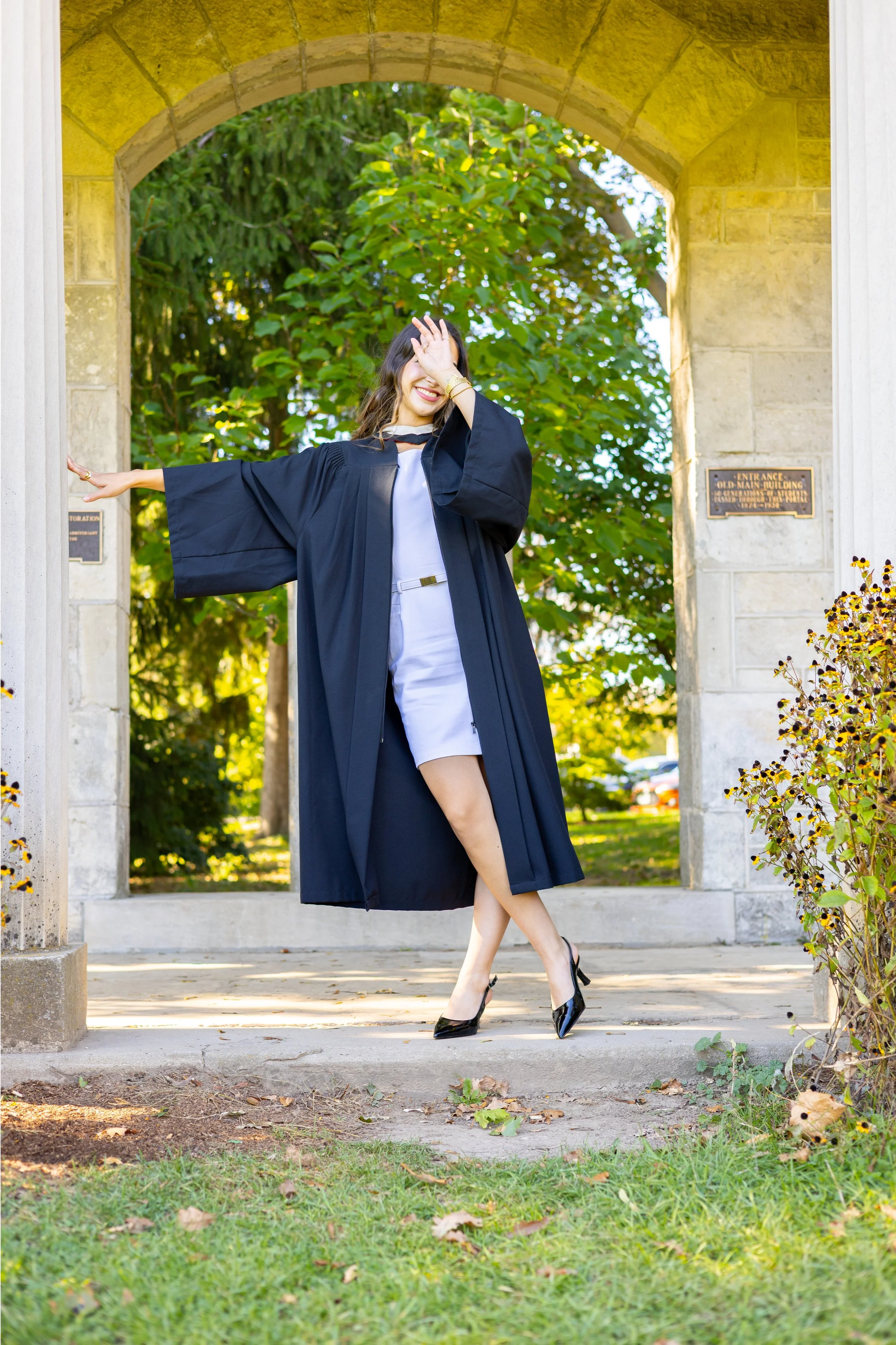 A young woman in a graduation gown and white dress, wearing black heels, standing under an archway with trees in the background, smiling and covering part of her face with her hand.