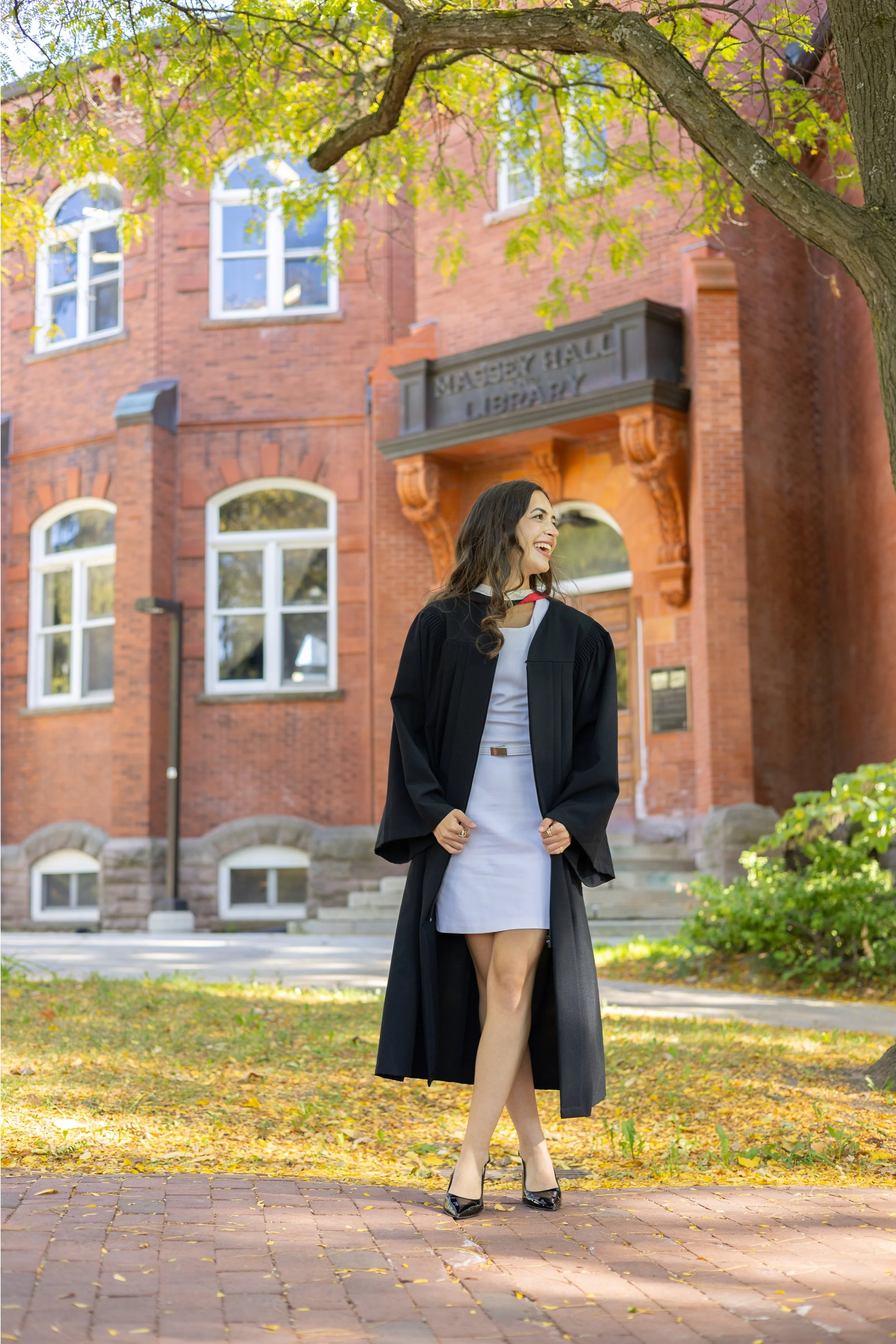 A young woman in a white dress and black graduation gown standing outdoors on a brick courtyard with fallen leaves, smiling and looking to her right, near a tree with green and pink leaves, in front of a brick building with arched windows and a sign 