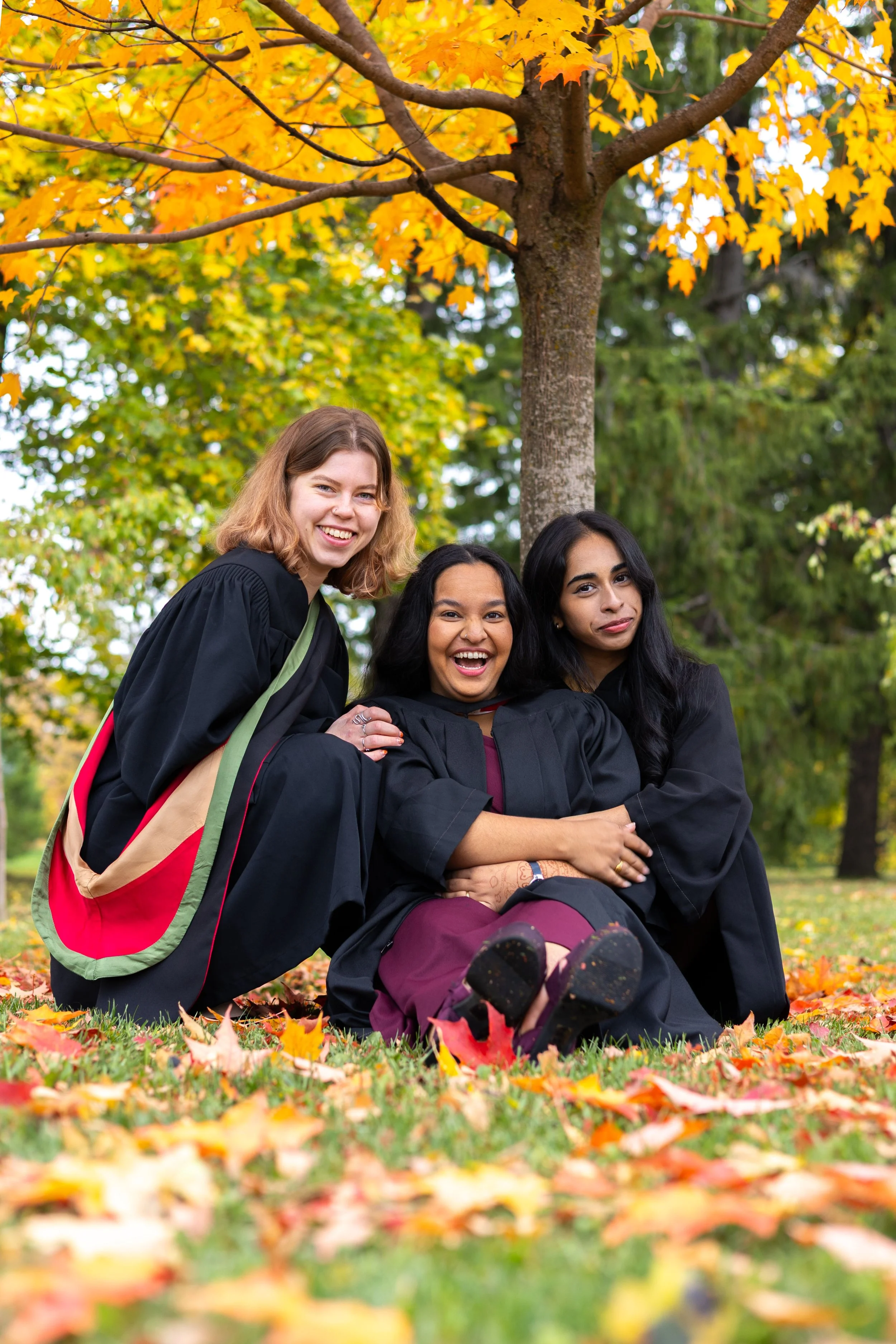 Three women in graduation caps and gowns sitting on fallen autumn leaves under a tree with yellow and orange foliage, smiling and embracing each other.
