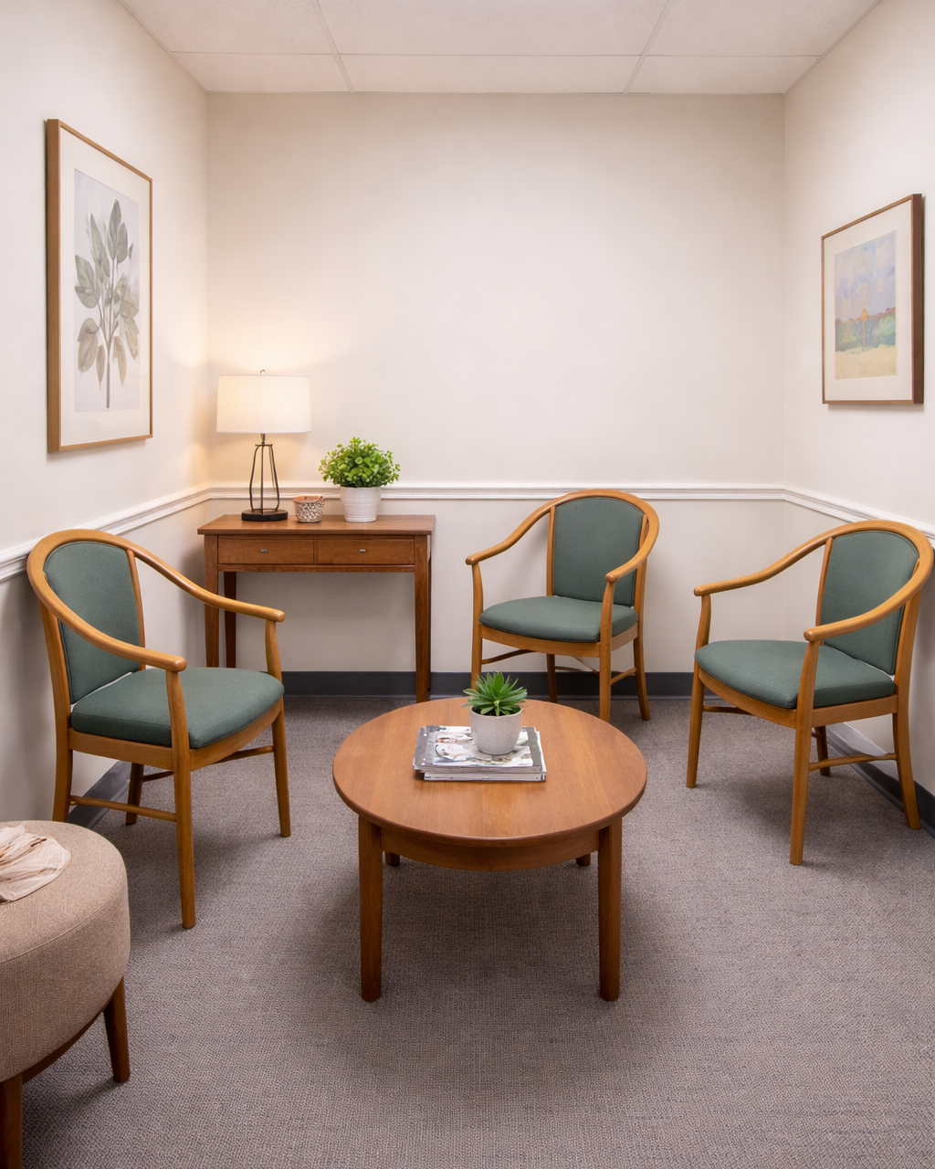 The Vitality Chiropractic and Wellness waiting room has upholstered chairs, a round wooden coffee table with books and a small potted plant, a small wooden side table with a lamp and a green plant, and framed artwork on the cream-colored walls.