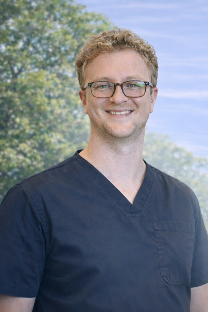 A man with blonde curly hair and glasses, wearing navy medical scrubs, standing outdoors with trees and a blue sky in the background.