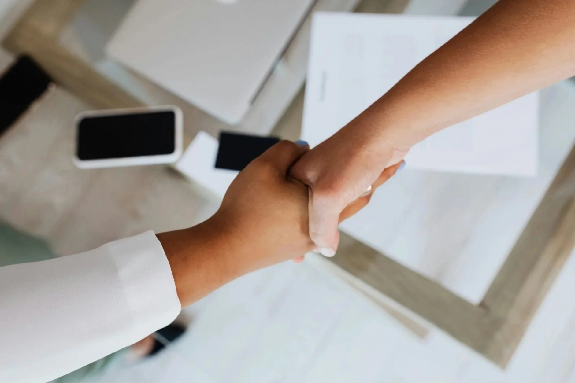 Two people shaking hands over a desk with a laptop and smartphones; relaying a partnership.