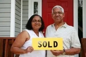 A woman and an older man standing in front of a house, holding a yellow sign that says SOLD. They are smiling.