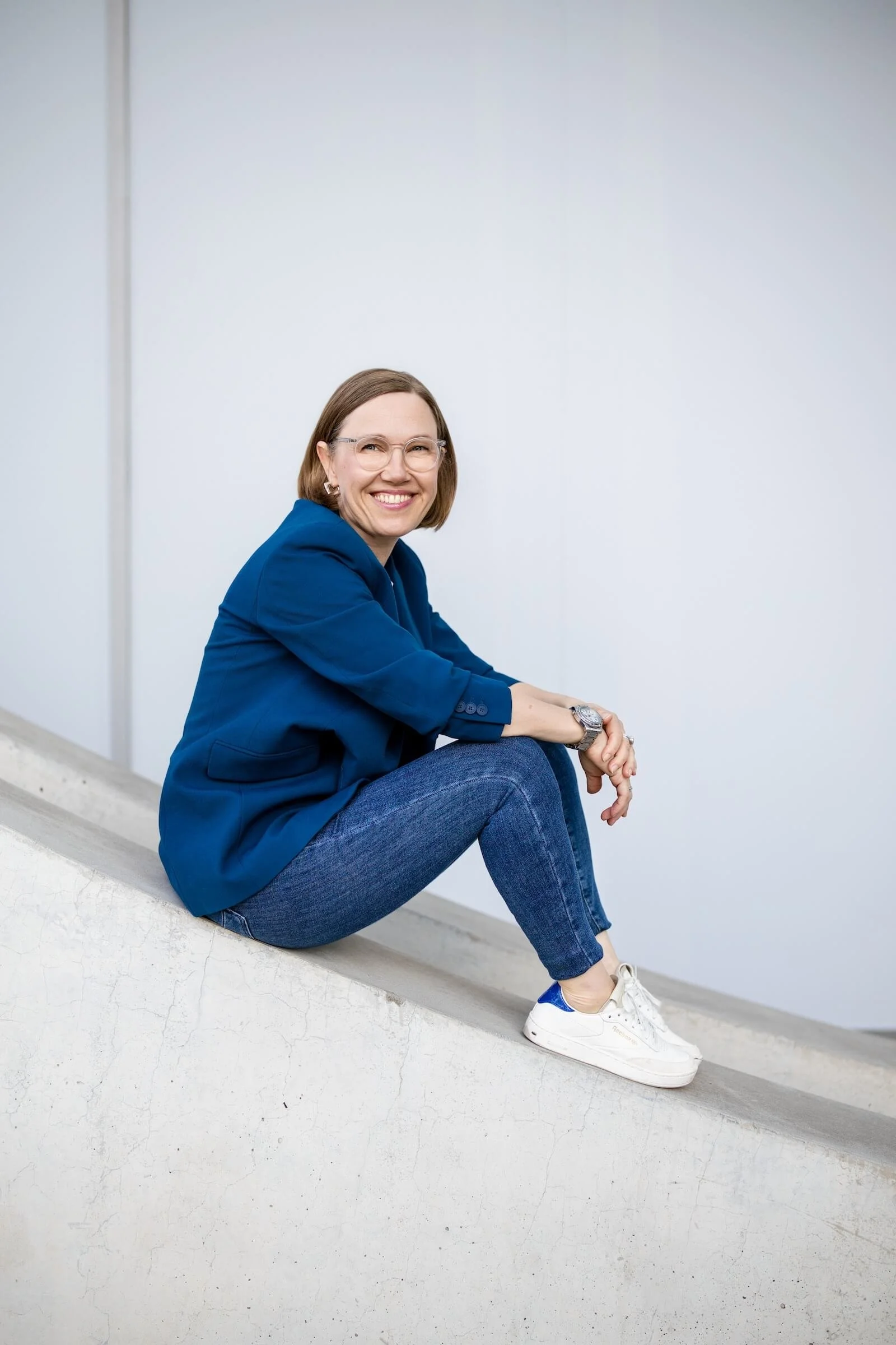 Eine lächelnde Frau mit Brille, blauer Blazer, Jeans und weißen Sneakern sitzt auf einer schrägen Betonfläche vor einem weißen Wand.