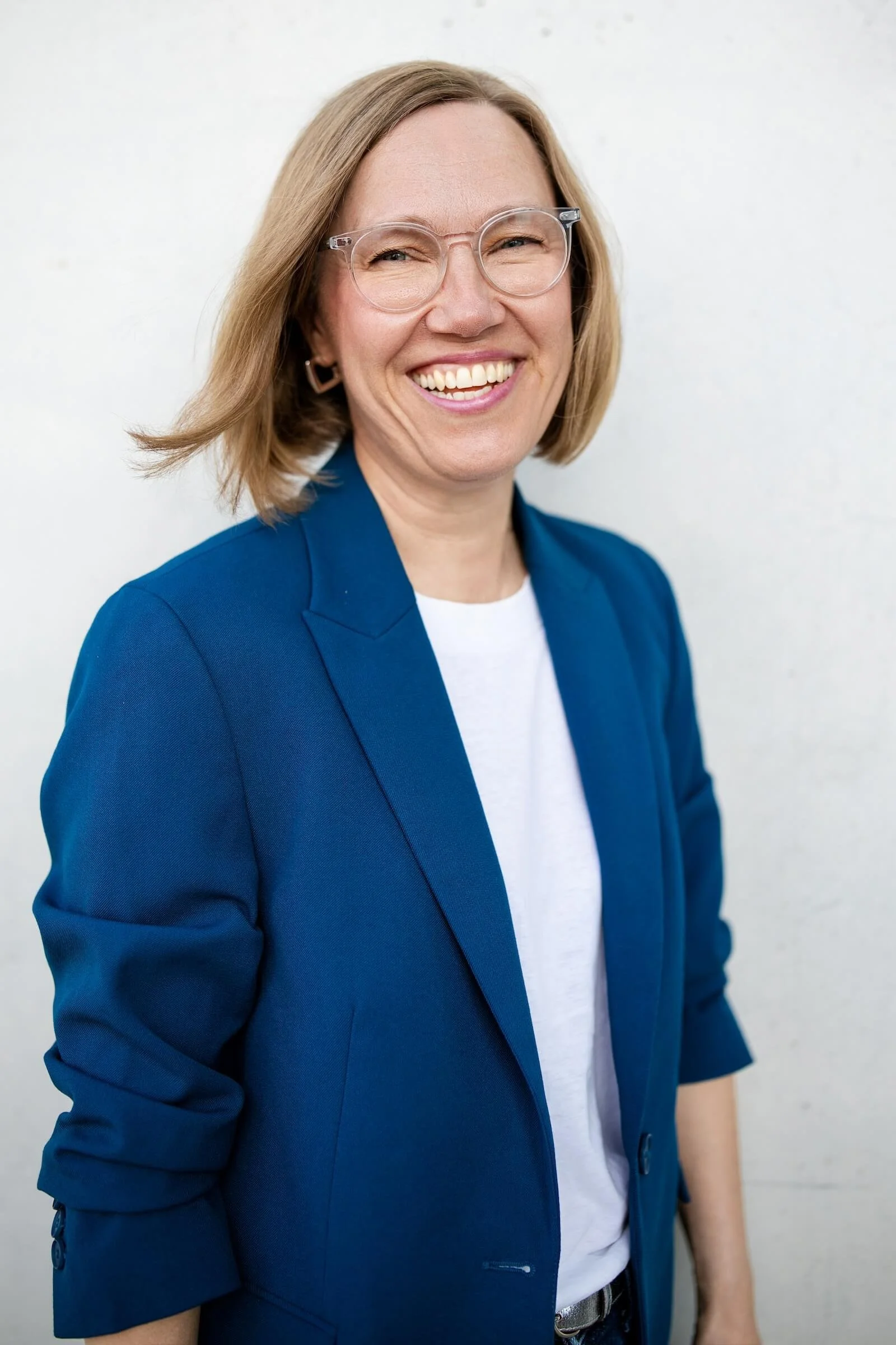 Eine lächelnde Frau mit Brille und blonden Haaren trägt eine blaue Blazer und ein weißes T-Shirt vor einem weißen Hintergrund.