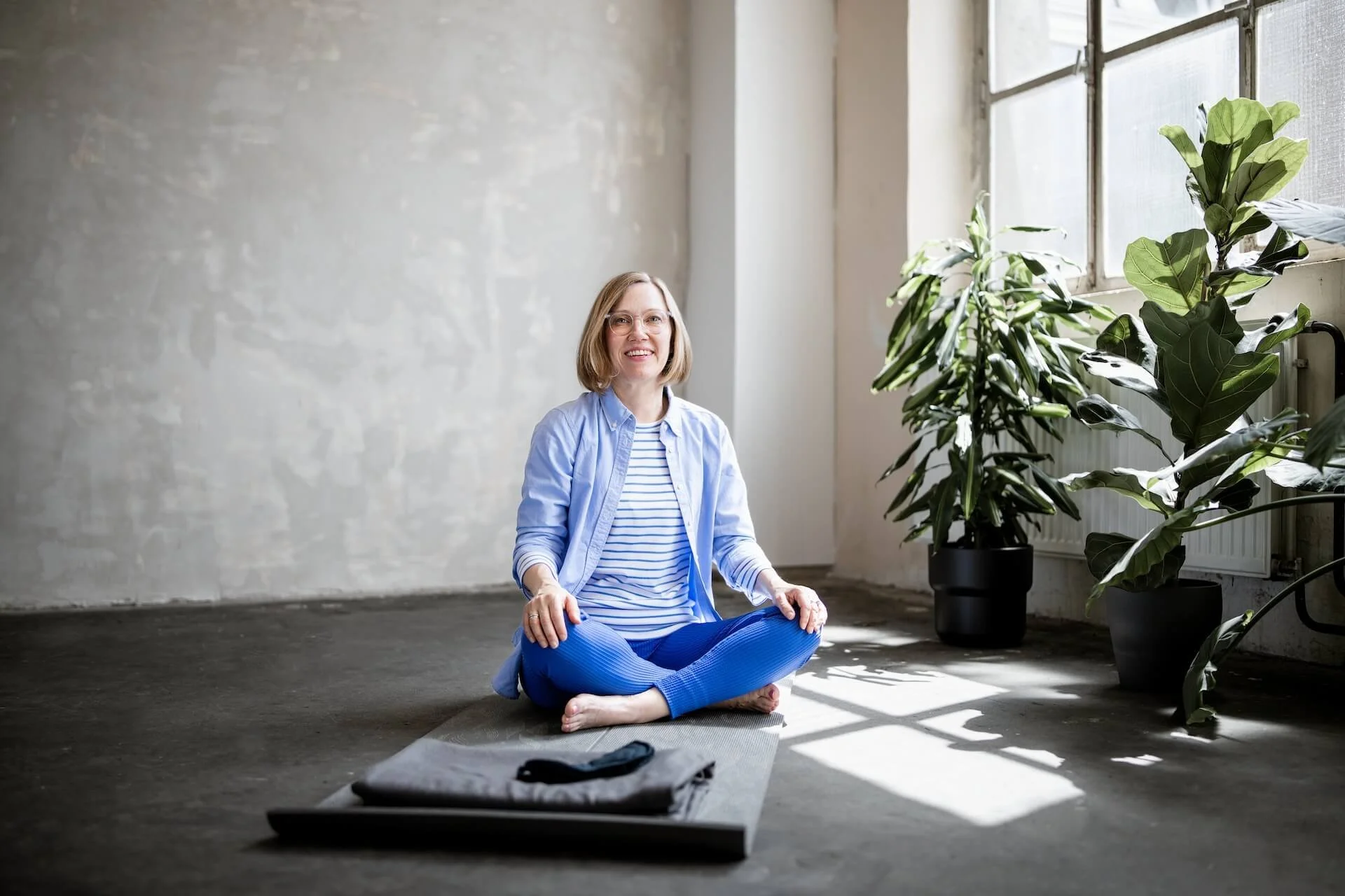 Eine Frau sitzt im Schneidersitz auf einer Yogamatte in einem Raum mit großen Fenstern und einigen Zimmerpflanzen.