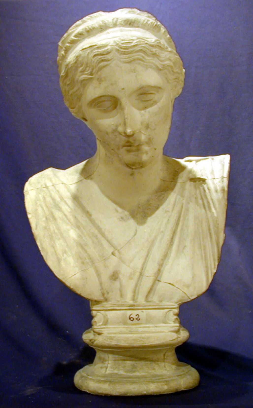 Busto de mármol de una mujer con cabello trenzado y ropa clásica, en un fondo azul.