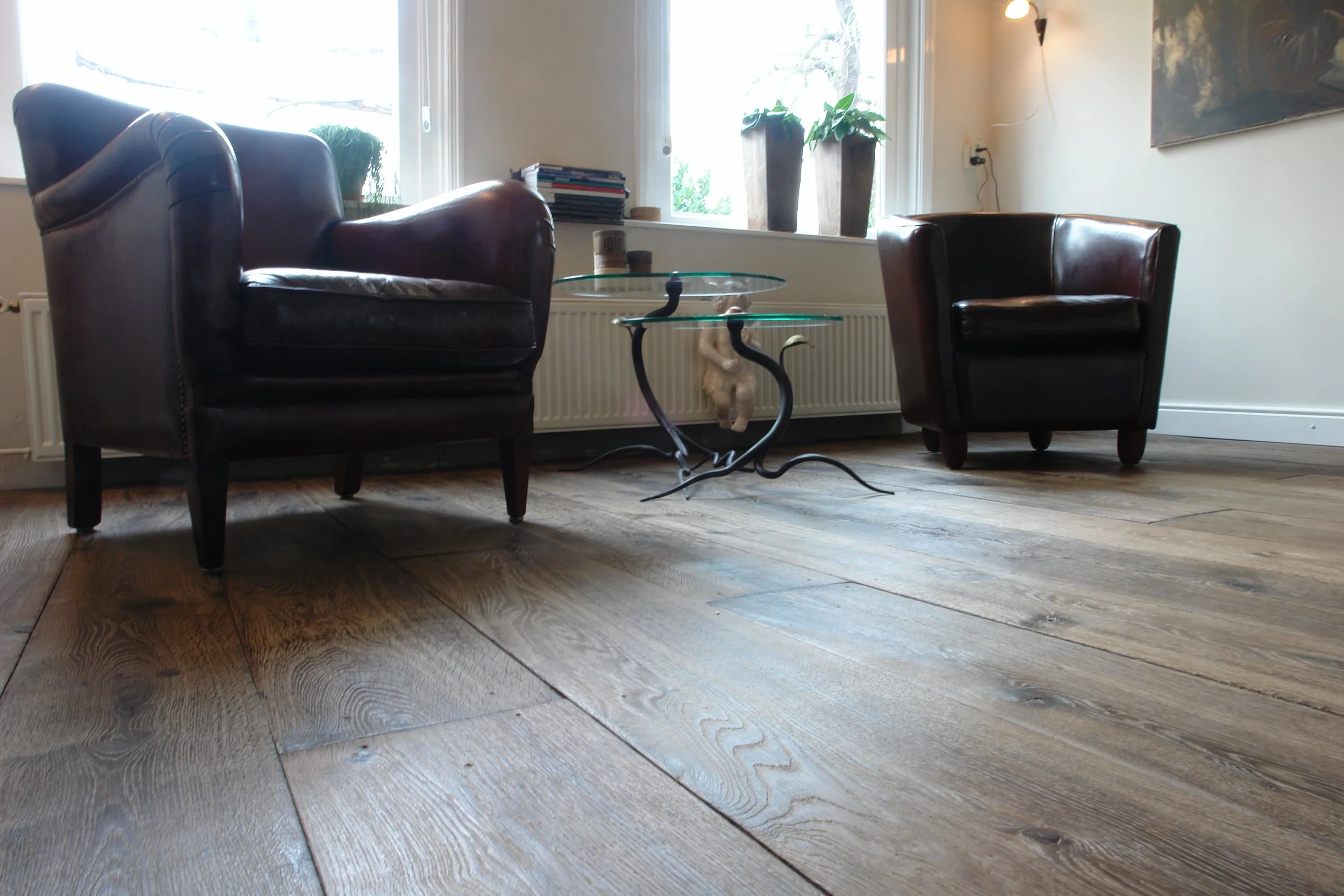 Sala con dos sillones de cuero oscuro y una mesa de vidrio con patas de hierro artístico, en una sala con ventanas y plantas.